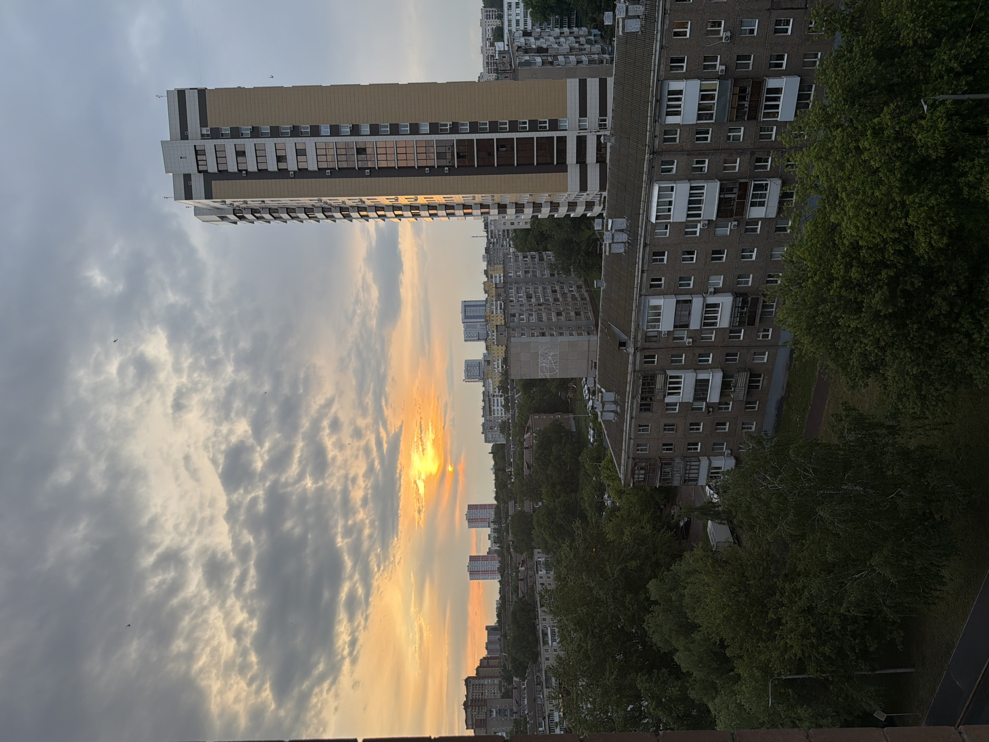 The image captures an urban sunset scene. Tall buildings dominate the view, interspersed with trees. The sky is filled with clouds tinted orange and grey by the setting sun. The picture appears to be taken from an elevated position, likely a high-rise building.