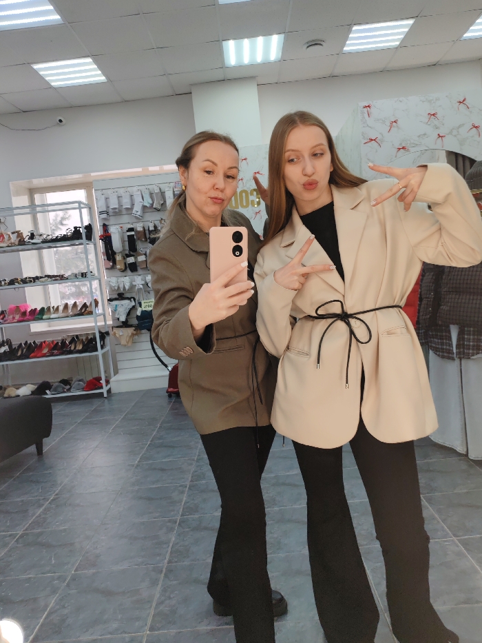 Two women pose for a selfie in a clothing store. The woman on the left holds a pink phone, wearing a khaki blazer and black pants. The woman on the right wears a cream-colored blazer with a black tie at the waist and black pants. She gestures with a peace sign. Racks of clothes and accessories line the background, adding to the store's ambiance. The room is well-lit with fluorescent lights.