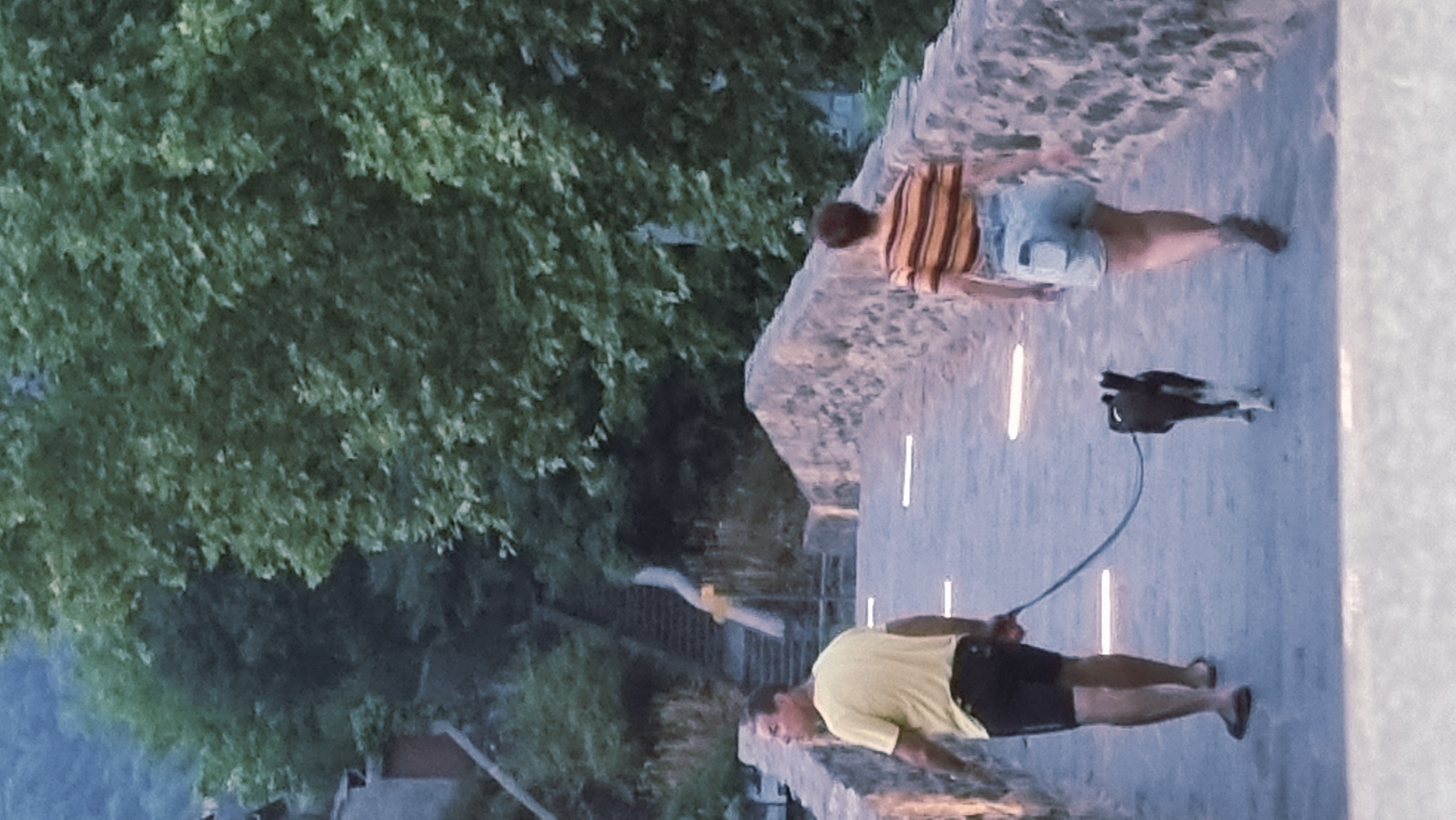 In Sant Joan Les Fonts, Spain, two individuals, a male and a female, walk across a stone bridge. The male, wearing a yellow t-shirt and black shorts, is leading a black and white dog on a leash. The female, in a striped t-shirt and denim shorts, walks ahead of the dog. The bridge is illuminated by several embedded lights, and a stone wall borders its edge. In the background, lush green trees and a stone staircase are visible.