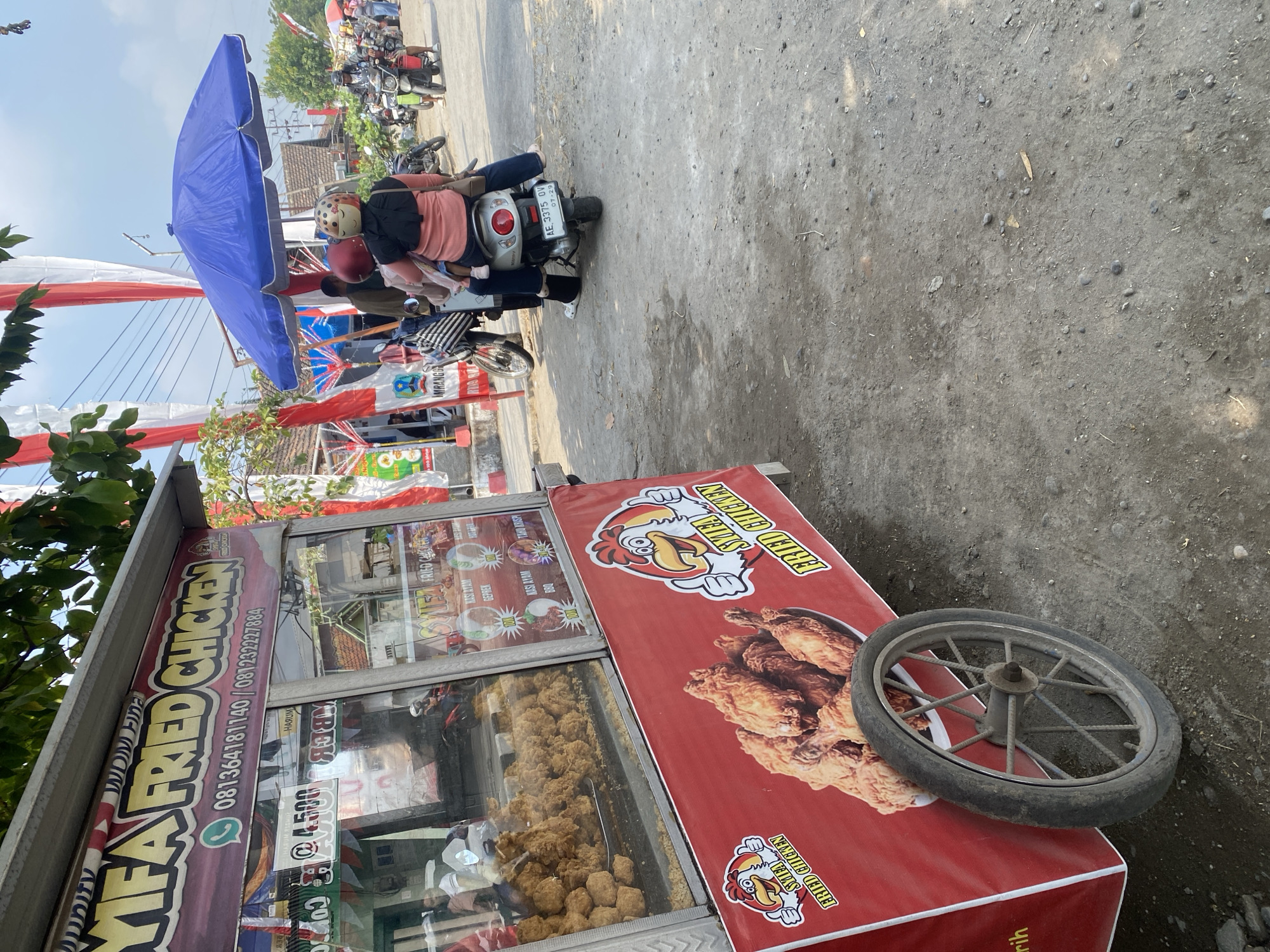 An outdoor scene in Sempol, Indonesia features a red fried chicken cart with branding that reads "SYIFA FRIED CHICKEN". The cart displays images of fried chicken and text in Indonesian. A glass display case on the cart showcases a large quantity of fried chicken. To the right of the cart, a blue umbrella is set up, shading a motorbike carrying two people, a woman driving and a child behind her. The woman is wearing a helmet adorned with a smiley face. In the background, other motorbikes and pedestrians are visible, along with buildings and red and white banners, suggesting a festive or local event. The ground is unpaved, consisting of dirt and small stones. The lighting indicates it is daytime.