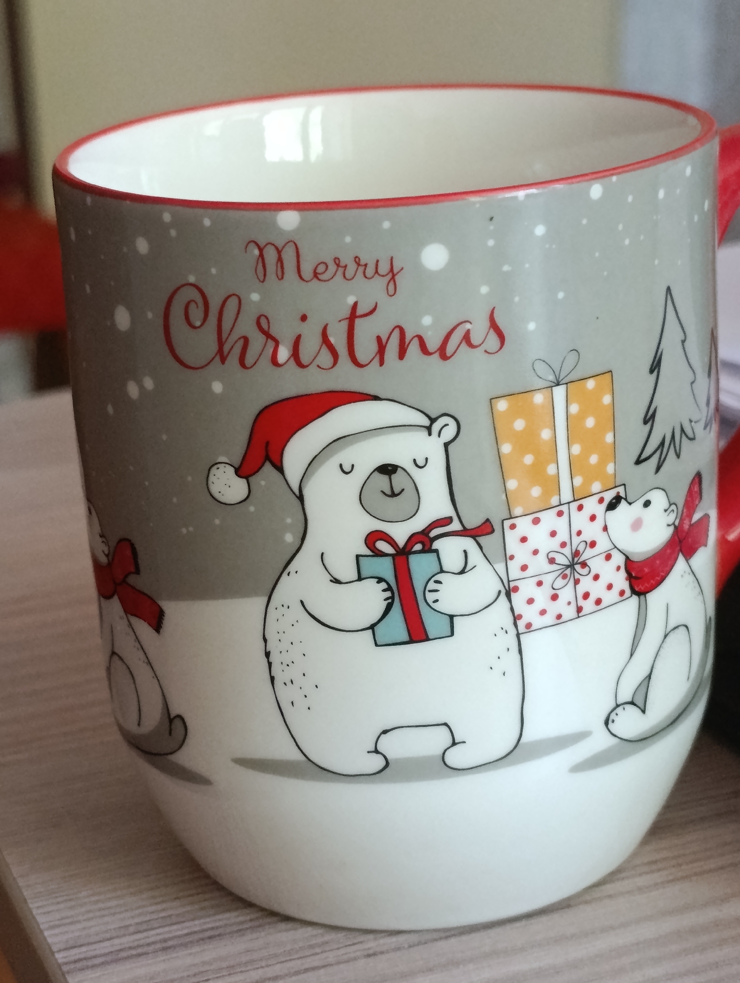 This is a close-up, eye-level, medium shot of a white ceramic mug with a gray background featuring a Christmas-themed illustration of polar bears and presents. The mug has a red rim and a red handle, partially visible on the right. The illustration depicts a large polar bear wearing a Santa hat, holding a light blue gift box with a red ribbon, and a smaller polar bear wearing a red scarf, looking up at a stack of two wrapped presents. The presents are wrapped in red and white polka-dot paper and orange paper with white polka dots. A simple black outline of a Christmas tree is visible in the background, along with scattered white dots representing snow. The mug sits on a light brown, horizontally-grained surface, likely a table. The background of the image is blurred, with hints of a red object and a neutral-toned wall.