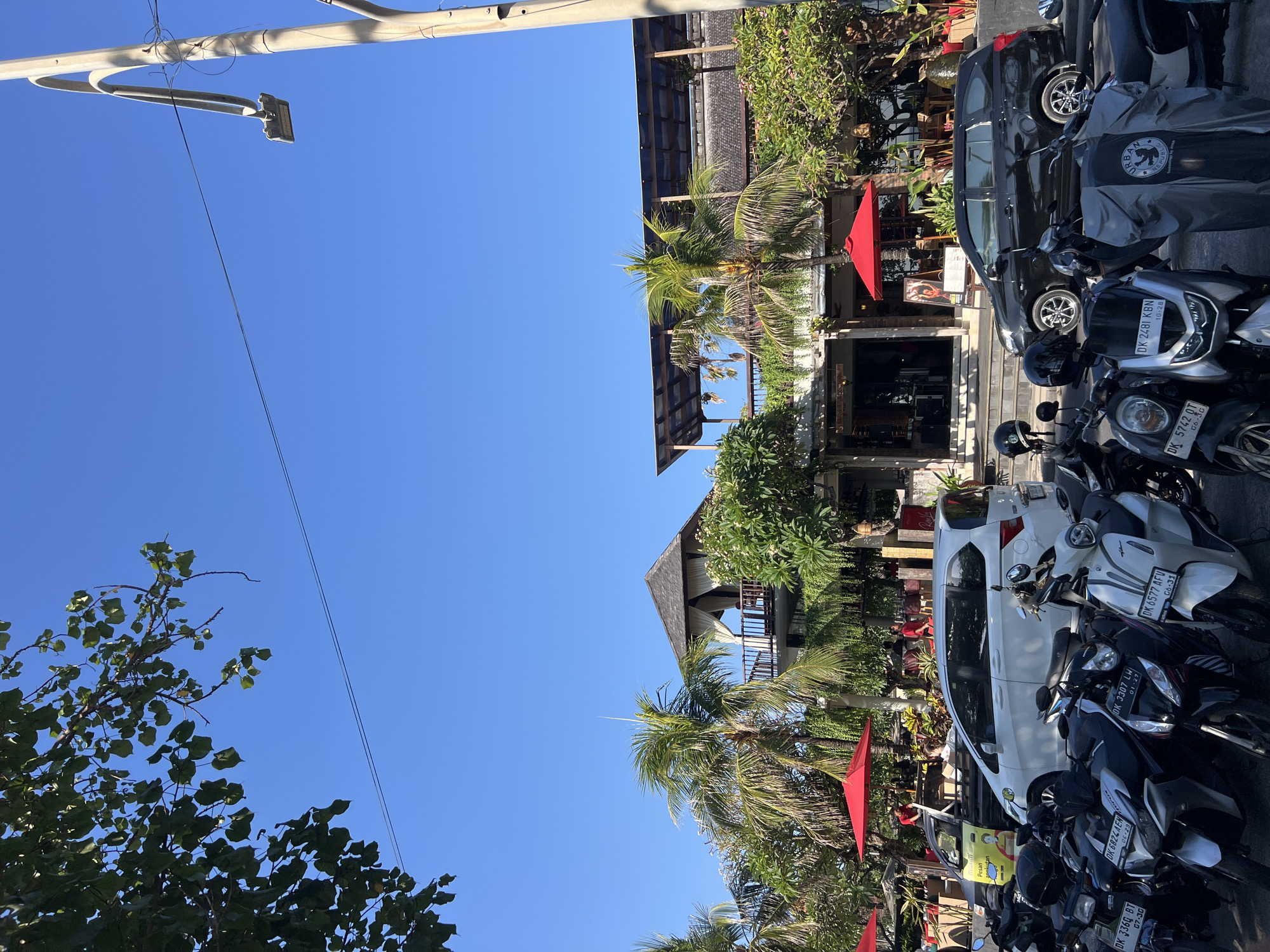 The image captures a street scene in Kuta, Indonesia, on a sunny day with a clear blue sky. In the foreground, a row of parked motorcycles, mostly scooters, are visible. These include a silver Honda with license plate DK 6577 AFV, a black Honda with license plate DK 3307 LWH, a white and black scooter with license plate DK 6824 EA, and a black scooter with license plate DK 336G BT. Some of the motorcycles are covered with gray tarps.
Behind the motorcycles, a white minivan is parked, and next to it, a black SUV. A yellow truck with a sign on its side is also present. The background features a multi-story building with a tropical architectural style, characterized by open-air balconies, wooden elements, and thatched roofs. Palm trees and lush green vegetation surround the building. Red umbrellas are set up on outdoor seating areas associated with the building, suggesting it is a restaurant or cafe. People are visible in the background, some seated at tables and others standing. The street appears to be paved. The overall impression is of a busy, sunny day in a tourist-oriented area.