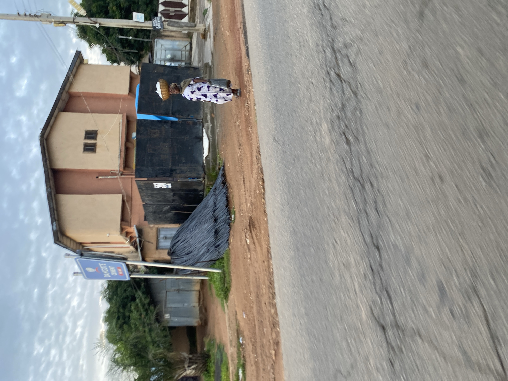 The image depicts a street scene in Jos, Nigeria, characterized by a weathered road in the foreground and a building with an attached structure in the background. A lone woman is walking on the right side of the road, carrying a basket of items on her head and a bag in her hand. She is wearing a patterned dress and a scarf. To the left of the road, a pile of dark rebar is visible, alongside a metal shed and a "Dangote Cement" signpost. The sky is overcast with clouds.