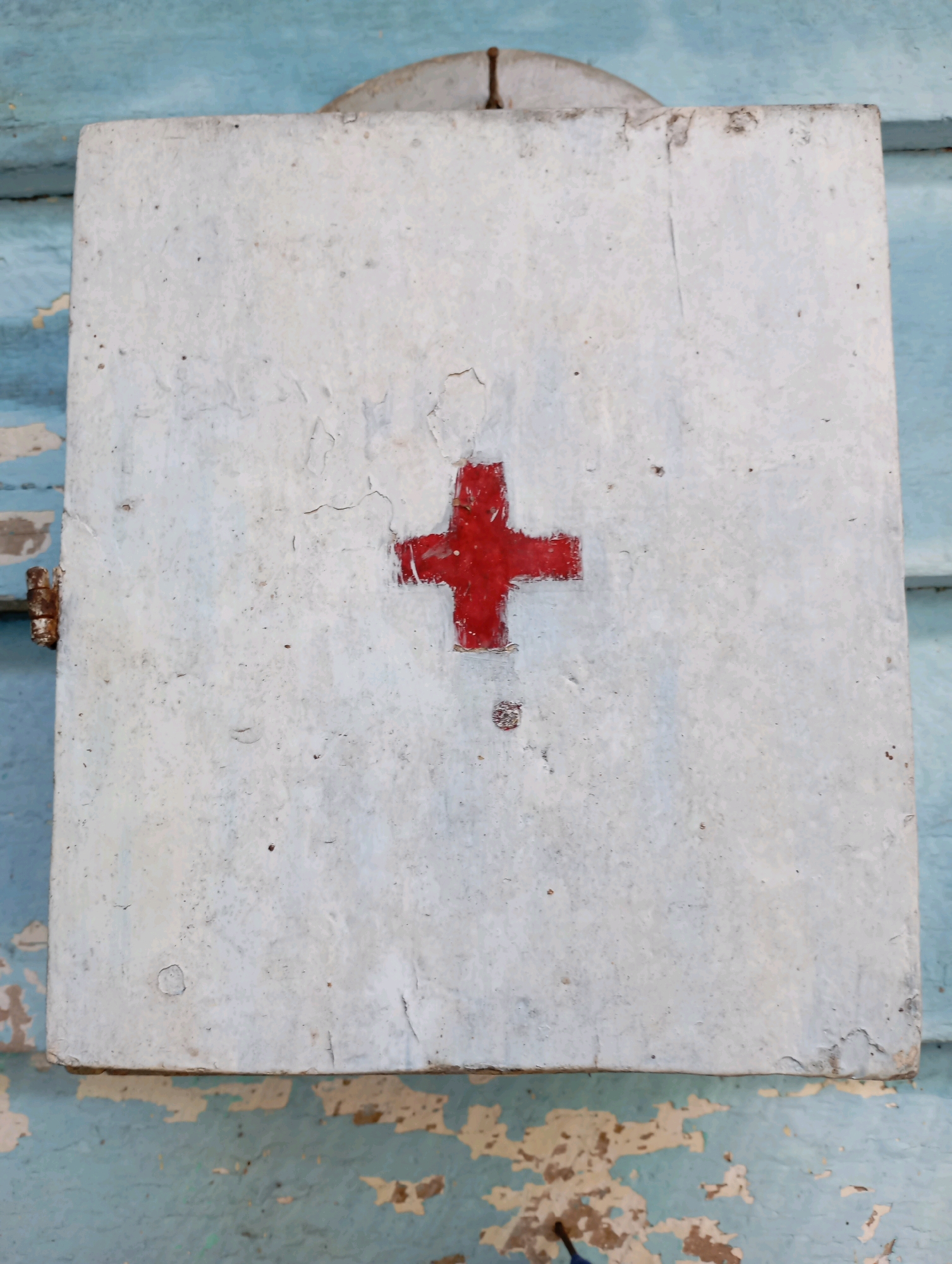 An aged, white, rectangular medical cabinet with a red cross painted on its front hangs on a weathered, light blue wooden wall. The cabinet appears to be made of metal and shows signs of rust around the hinge on the left side. The paint on the cabinet is chipped and peeling, revealing a grayer undertone in places. The red cross, though still visible, also exhibits wear and fading. The wooden wall behind the cabinet is also in poor condition, with large sections of paint missing, exposing the natural wood beneath. The scene suggests an outdoor location, possibly in a disused or rustic area. The image does not contain any people or provide any specific clues about its exact location within Balikpapan, Indonesia, other than the general context of an urban environment in that city. The overall impression is one of neglect and the passage of time.