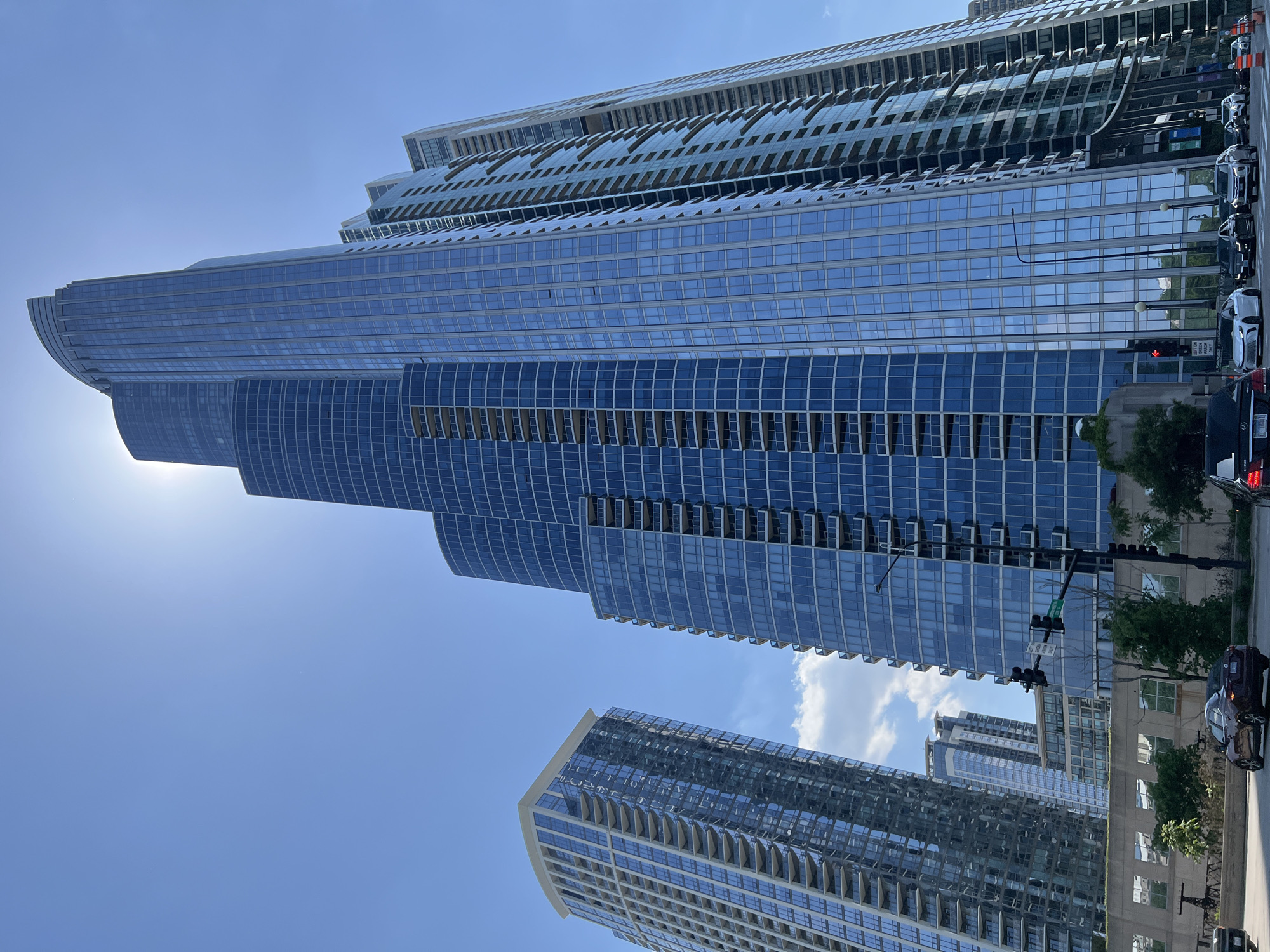 The image shows a low-angle view of several modern skyscrapers in an urban environment. The primary building features a distinctive, stepped design with a curved facade and a predominantly blue glass exterior. Other buildings, visible in the foreground and background, exhibit similar modern architectural styles with glass and metal facades. A street runs alongside the buildings, with parked cars, traffic signals, and some greenery (trees) visible. The sky is predominantly blue with a few scattered clouds.