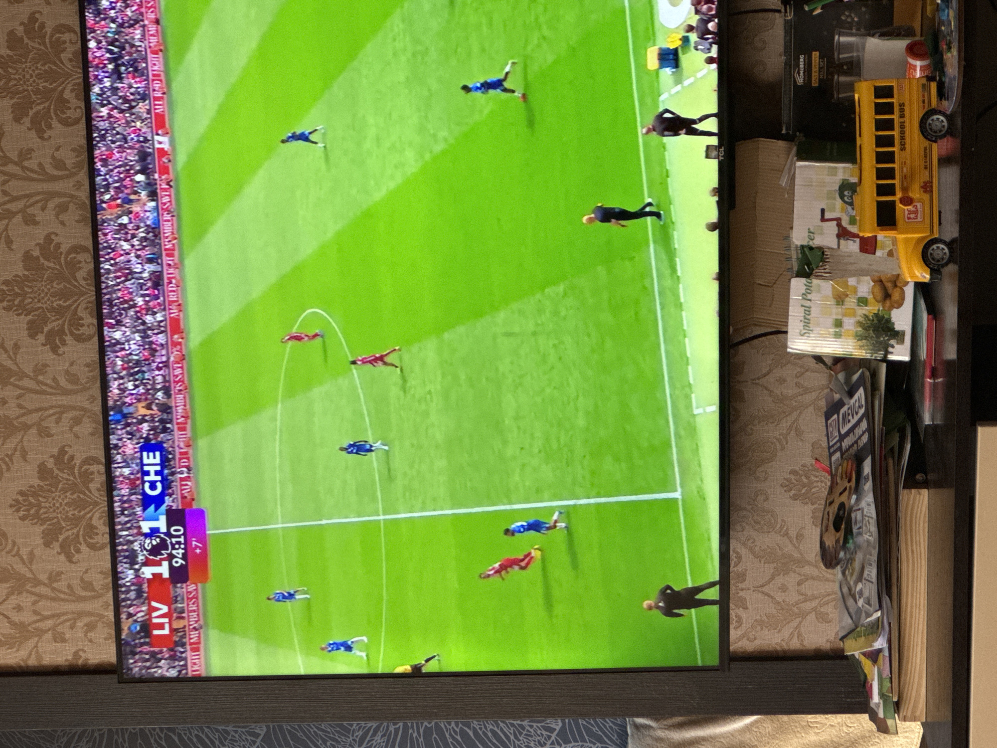 A television screen displays a soccer match with Liverpool (LIV) versus Chelsea (CHE). The score is tied 1-1 at 94:10 with +7 minutes of extra time. A green soccer field is visible on the screen with players in red and blue uniforms. The TV is surrounded by wallpaper with a floral design. To the right of the screen, there are assorted objects including a yellow toy school bus and boxes. The exact location in Yerevan, Armenia, cannot be determined from the provided image.