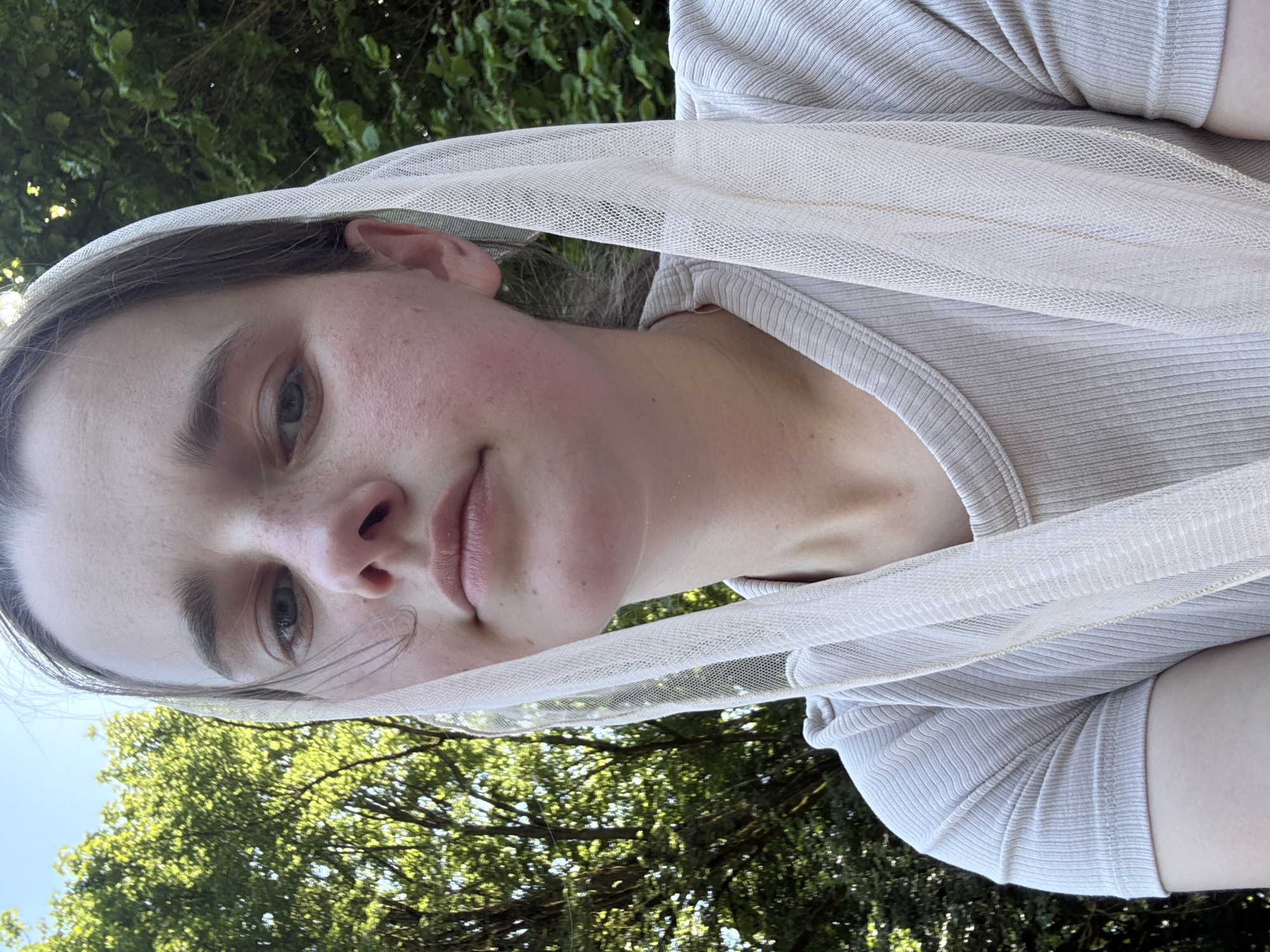 A young woman is photographed from the chest up, outdoors. She is wearing a light-colored, ribbed t-shirt and a sheer, white veil that drapes over her head and shoulders. Her face is partially obscured by the veil. She has light skin, light brown hair, and freckles across her nose and cheeks. Her eyes are blue-green. She is looking directly at the camera with a neutral expression. The background is out of focus, showing green foliage and a bright sky, suggesting a natural or park-like setting. The lighting indicates it is daytime. The location is Beltershain, Germany.