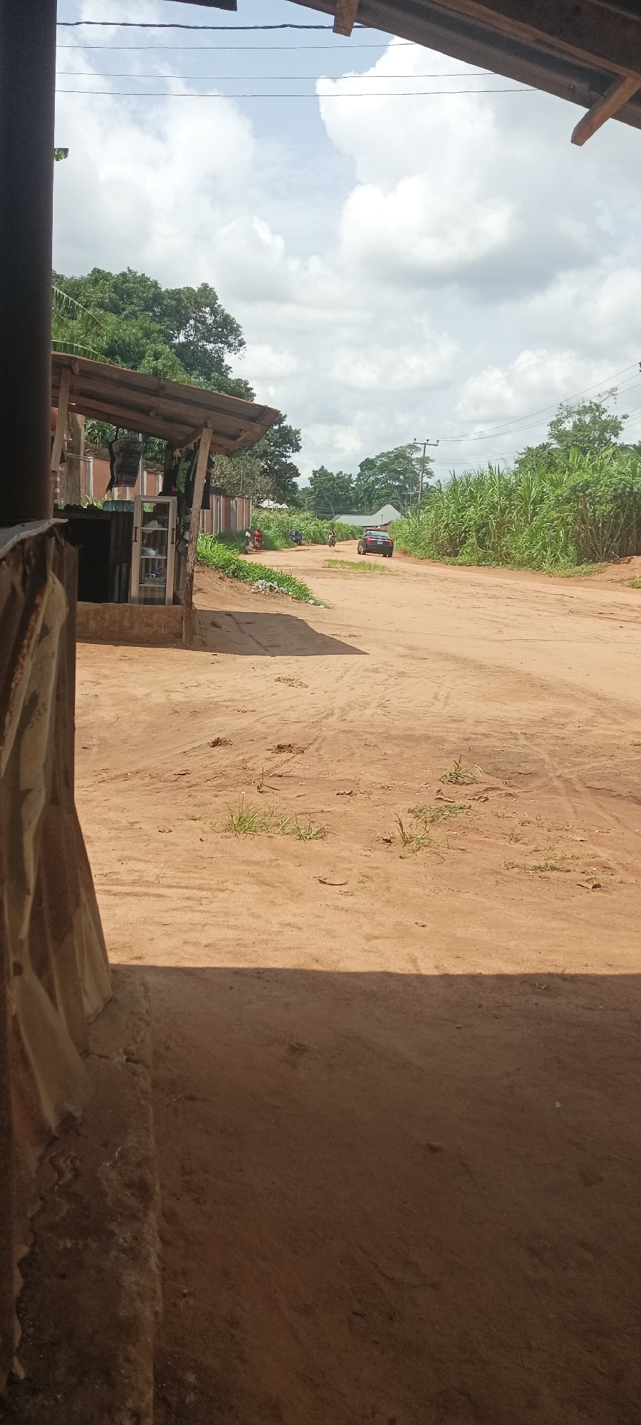 The image depicts a dirt road in Amauzu, Nigeria. A vehicle is visible in the distance on the road. The foreground shows a shaded area under a simple structure. There is vegetation on the sides of the road. In the distance, a building and power lines can be seen.