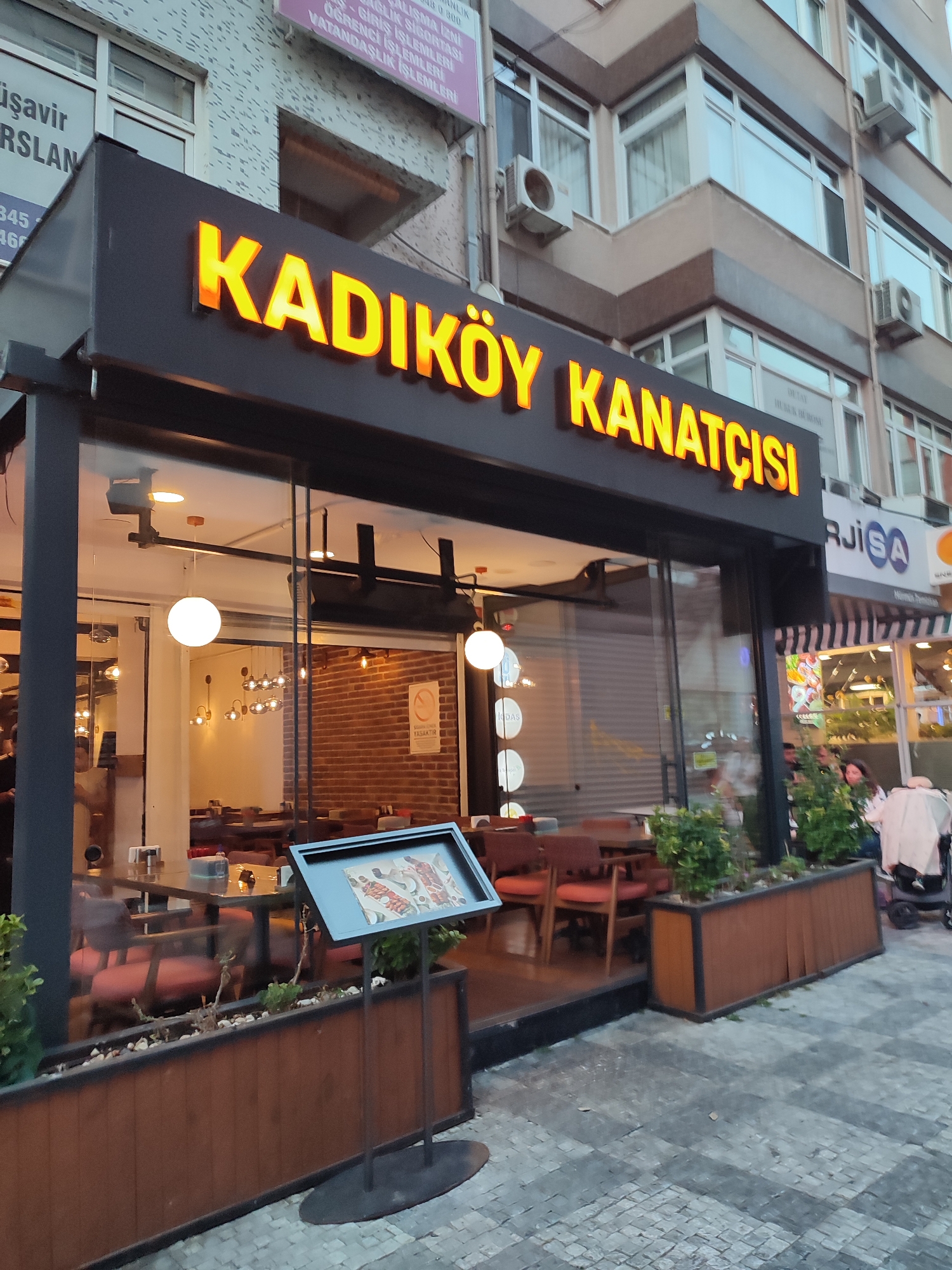 The image shows the exterior of a restaurant named "KADIKÖY KANATÇISI" in Kadıköy, Turkey. The restaurant has a modern facade with large glass windows revealing the interior, which is lit by pendant lights and features brick walls. The sign for the restaurant is illuminated with bright orange letters. In front of the restaurant, there are planters with greenery and a display stand with a menu. The street outside is paved with cobblestones. A few people are visible near the entrance of the restaurant, including a woman pushing a stroller. The overall scene depicts a commercial area in a city, likely in the evening or early morning given the lighting.