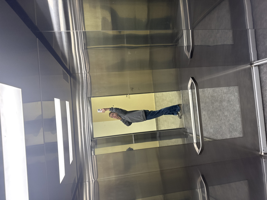 An image depicts the interior of an elevator car, located in Unknown, Unknown. The elevator features brushed metallic walls, a grey tiled floor, and a reflective silver ceiling with two prominent rectangular fluorescent light fixtures. In the center, the highly reflective elevator doors show the full-body reflection of a single individual, identified as an adult female. She is wearing a grey long-sleeved top, blue jeans, and light-colored slip-on shoes. Her left hand is raised, holding a white smartphone towards her reflection, indicative of taking a selfie. Her right hand is positioned near her chin. Her head is tilted slightly, and her gaze is directed towards the smartphone screen within the reflection. Two polished metal handrails are mounted horizontally across the lower sections of the elevator door panels. Beyond the reflected doors, a hallway with a light yellow wall and a grey floor is partially visible.