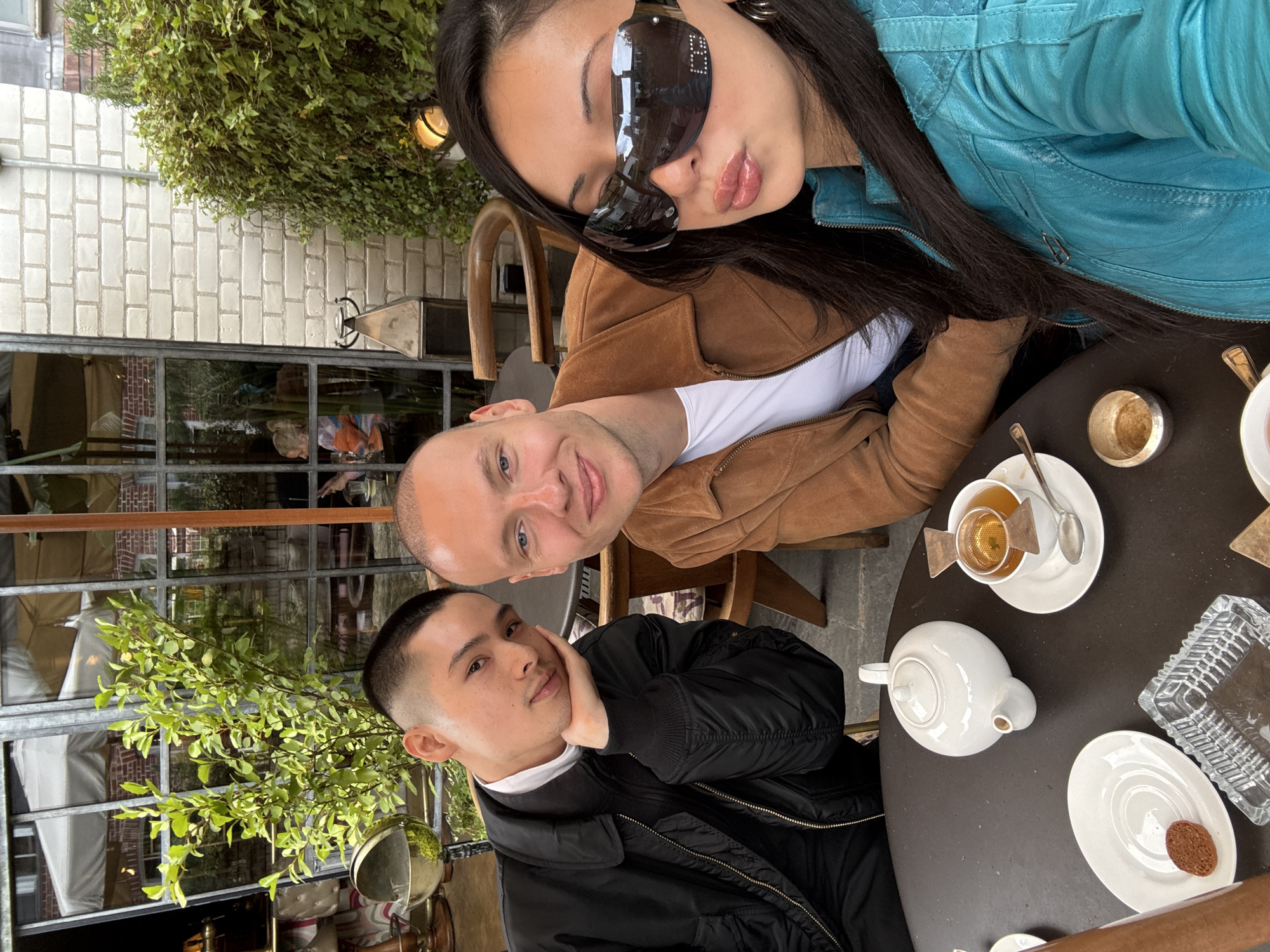 Three individuals, two males and one female, are captured in what appears to be a selfie taken at an outdoor cafe or restaurant. The image is rotated clockwise, making the subjects appear on their sides.
The female is on the right side of the frame, wearing large, dark rectangular sunglasses that obscure her eyes. She has long, dark hair and is wearing a bright teal or turquoise quilted jacket over a brown top. Her lips are pursed in a slight pout.
In the middle is a male with a bald head, dressed in a brown jacket over a white t-shirt. He is looking towards the camera with a friendly smile.
On the left side of the frame is another male with a neat, short dark haircut (fade). He is wearing a black bomber jacket over a white-collared shirt and is resting his chin on his hand. He looks calmly at the camera with a subtle, slight smile.
They are seated around a dark, round table. On the table, there is a white teapot, a white teacup containing tea with a metal infuser, a saucer with a small brown biscuit, an empty golden metallic cup (possibly an ashtray or condiment holder), and a clear glass ashtray.
The background features a building with light-colored brick walls, large windows reflecting the surroundings, and lush green foliage, possibly ivy or a hanging plant, cascading over a section of the wall. A light fixture is visible on the brick wall.
The activity appears to be a casual social gathering, possibly over tea, with the group posing for a photograph. The lighting suggests it is daytime, though the exact time is unclear. The overall mood seems relaxed and convivial. No visible text is present in the image. The location context provided indicates this scene is in London, United Kingdom.