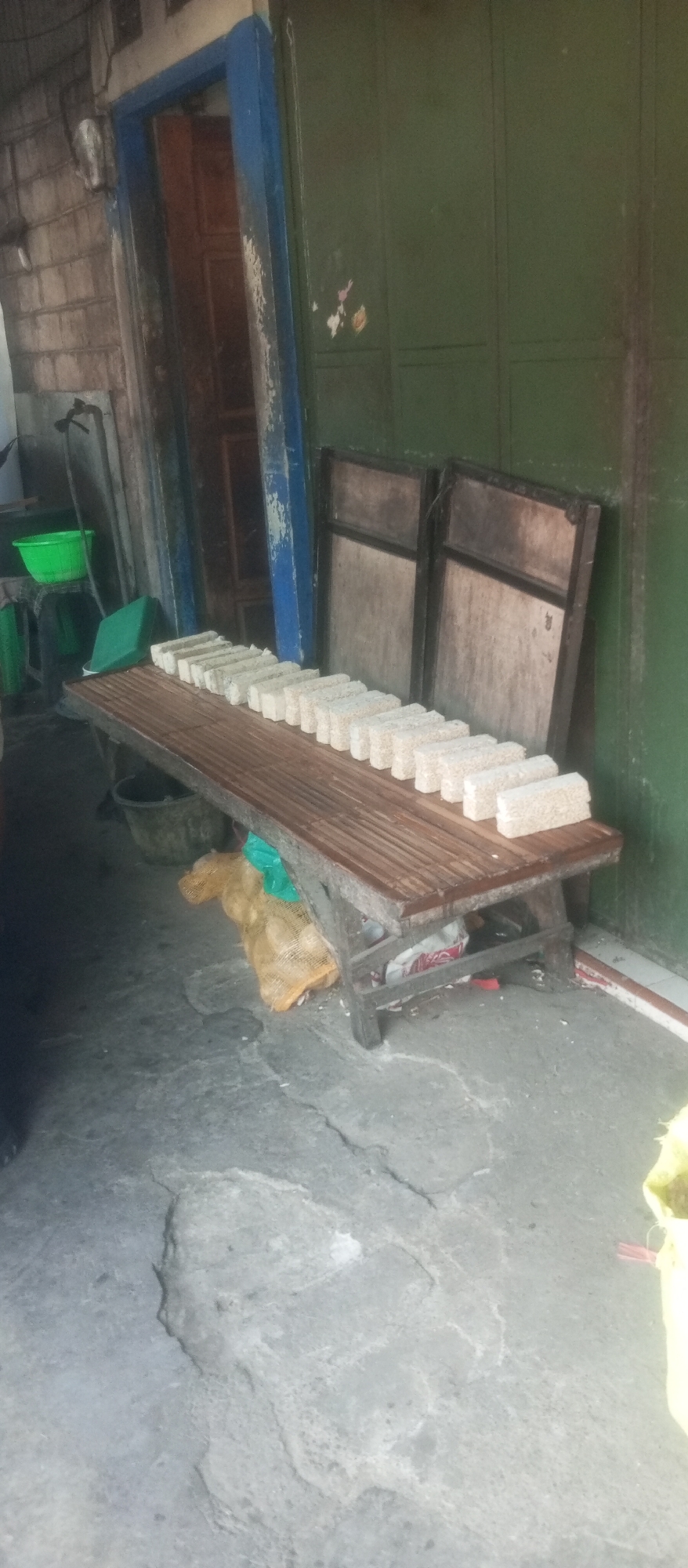 The image shows an outdoor scene, possibly a market or a processing area, with a row of rectangular, white objects neatly arranged on a wooden bench. The bench appears to be made of bamboo slats and has a rustic, aged look. Behind the bench, there is a blue-framed doorway leading into a dimly lit interior. To the right of the doorway, a large, dark green metal door is visible, with some peeling paint and what appears to be stickers on its surface. The ground is concrete and shows signs of wear and tear, with visible cracks. To the left of the bench, a green plastic bucket sits on a stand, and behind it, a dark hose or pipe is visible. Underneath the bench, a sack filled with what looks like produce, possibly potatoes or onions, is placed on the ground, partially covered by a green plastic sheet. The overall impression is of a functional, utilitarian space. The presence of the food items and the rustic setting suggest a possible location in or near a local market or food production area within Kediri City, Indonesia, where such goods are commonly sold or processed.