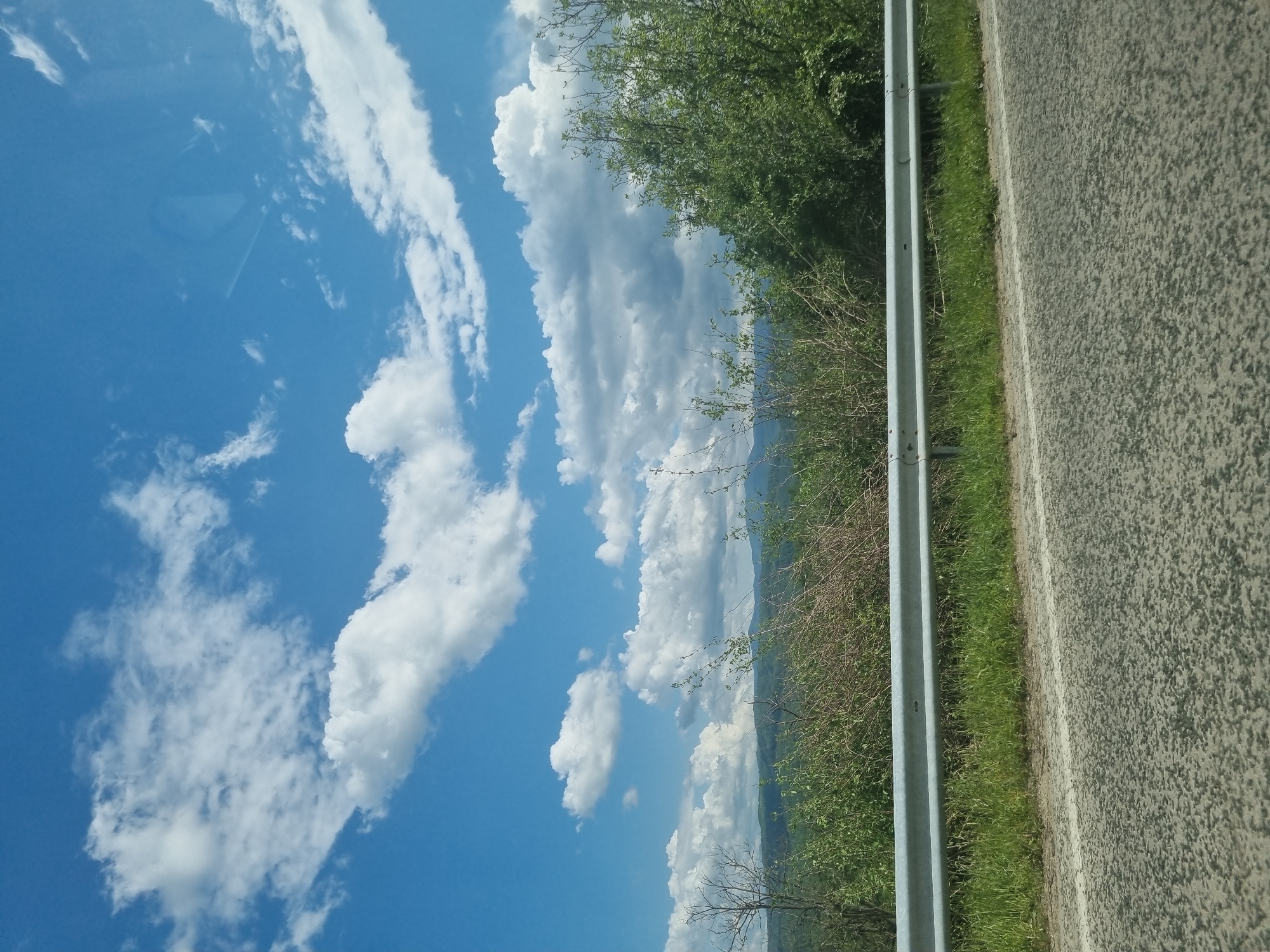 A roadside scene, possibly in or near Брусовeте, Bulgaria, shows a view of a blue sky with white clouds, roadside vegetation, a metal guardrail, and a paved road. The sky dominates the left side of the image, while the road and adjacent vegetation occupy the right side. The guardrail runs vertically between the vegetation and the road.