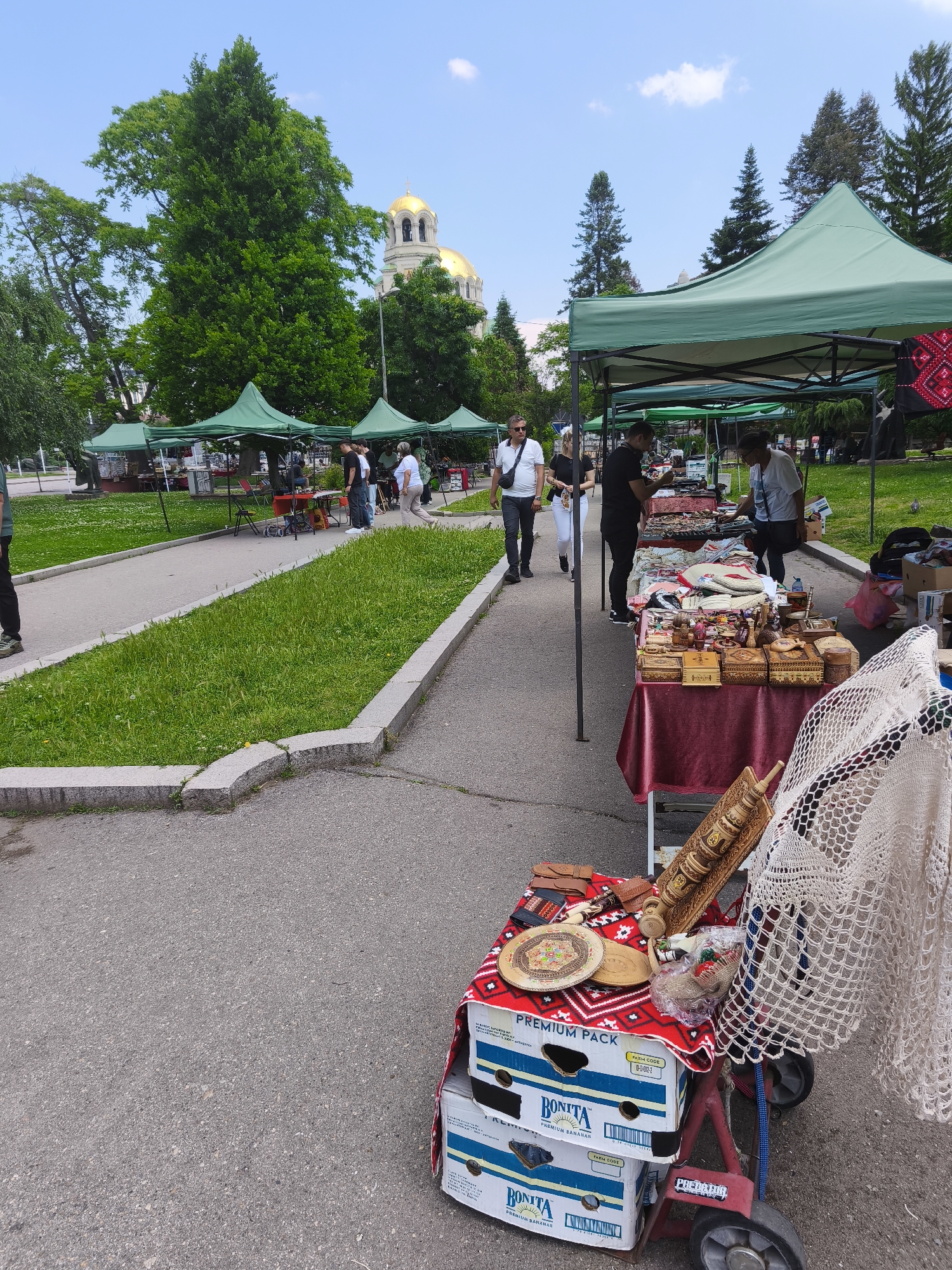 This image captures an outdoor market or fair situated on an asphalt path within a park in Sofia, Bulgaria. The prominent golden domes of the Alexander Nevsky Cathedral are visible in the background, partially obscured by tall, lush green trees, including both coniferous and broadleaf species. The sky is clear and blue with minimal white cloud cover.
The scene focuses on several green canopy tents set up along the path, housing various vendor stalls. In the foreground on the right, a table draped with a maroon cloth and a red and white patterned textile displays an array of handcrafted goods. These items include multiple carved wooden boxes, decorative wooden plates, a distinctive carved wooden cylinder, and various smaller trinkets. A large white crocheted textile hangs from the front of this display. Below the table, two stacked cardboard boxes labeled "Bonita Premium Bananas" rest on a red hand truck with "PREDATOR" branding, serving as risers for the merchandise.
Multiple individuals are present on the asphalt path. In the mid-ground, a man wearing a white polo shirt and sunglasses, with a black shoulder bag, walks from left to right. Behind him, a woman in a white top and light trousers also walks in the same direction. At the rightmost market stall, two individuals, one in a dark shirt and another in a white shirt, are standing and observing the items on display. The lower torso and legs of a person wearing green trousers and light-colored shoes are visible in the far-left foreground. The path is delineated by concrete curbs, bordering strips of green grass. The environment is well-lit by daylight, indicating a sunny or partially sunny day.
