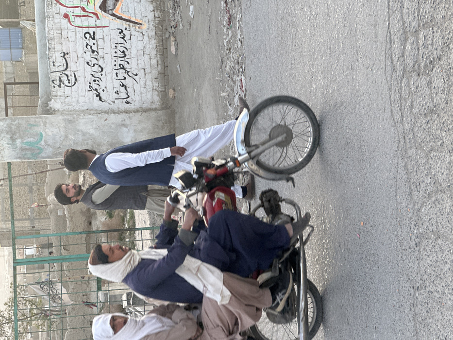This image depicts an outdoor urban scene in Quetta City Tehsil, Pakistan. The foreground features a paved asphalt road under natural daylight. On the left, a red motorcycle with chrome accents, including its front fender and exhaust pipe, is in motion. Two adult males are riding it: the driver, wearing a dark blue shalwar kameez and a white head covering, is positioned forward, and a passenger, dressed in a light brown shalwar kameez with a white head covering, is seated directly behind him. Both individuals are facing towards the right side of the frame relative to their direction of travel.
On the right side of the frame, a low, white-painted brick wall displays black and orange graffiti. The black script, written in Urdu, includes phrases such as "2 January, Friday" and indications for prayer times, specifically "after Zuhr prayer until Isha." Adjacent to this wall, two adult males are present on a dirt and debris-strewn shoulder. One man, in the midground, wears a dark gray vest over a light-colored shirt and tan shalwar, and is looking towards the left. The second man, positioned slightly behind him, wears a dark blue vest over a white shirt and white shalwar, with his back partially to the viewer as he appears to be walking towards the right.
In the background, on the left, a green metal fence partially obscures an area with earthen mounds and what appears to be a white flag or banner with dark script, suggestive of a cemetery or undeveloped land. Distant structures are visible on the horizon under a light sky.