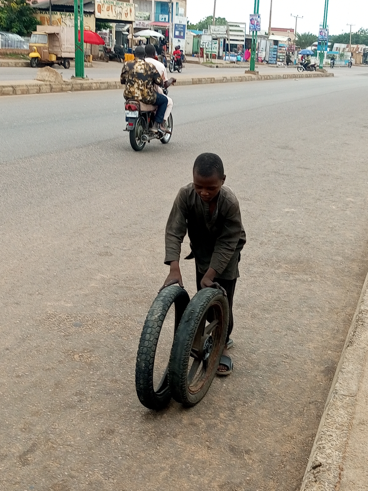 In Gombe, Nigeria, on a paved road in front of a row of shops, a young boy walks while carrying two motorcycle tires. He is wearing a dark grey long-sleeved shirt and black pants. The boy is on the right side of the frame, and the tires he is carrying are in front of him, held by his hands. Behind him, two men ride on a motorcycle. The man in front is wearing a patterned shirt, and the man behind him is wearing a light-colored shirt. The motorcycle has a visible license plate. The background shows a street with several small shops, some with signs. A yellow motorized tricycle is parked to the left. The road is grey asphalt, and there is a concrete curb on the right side of the frame. The lighting suggests it is daytime.