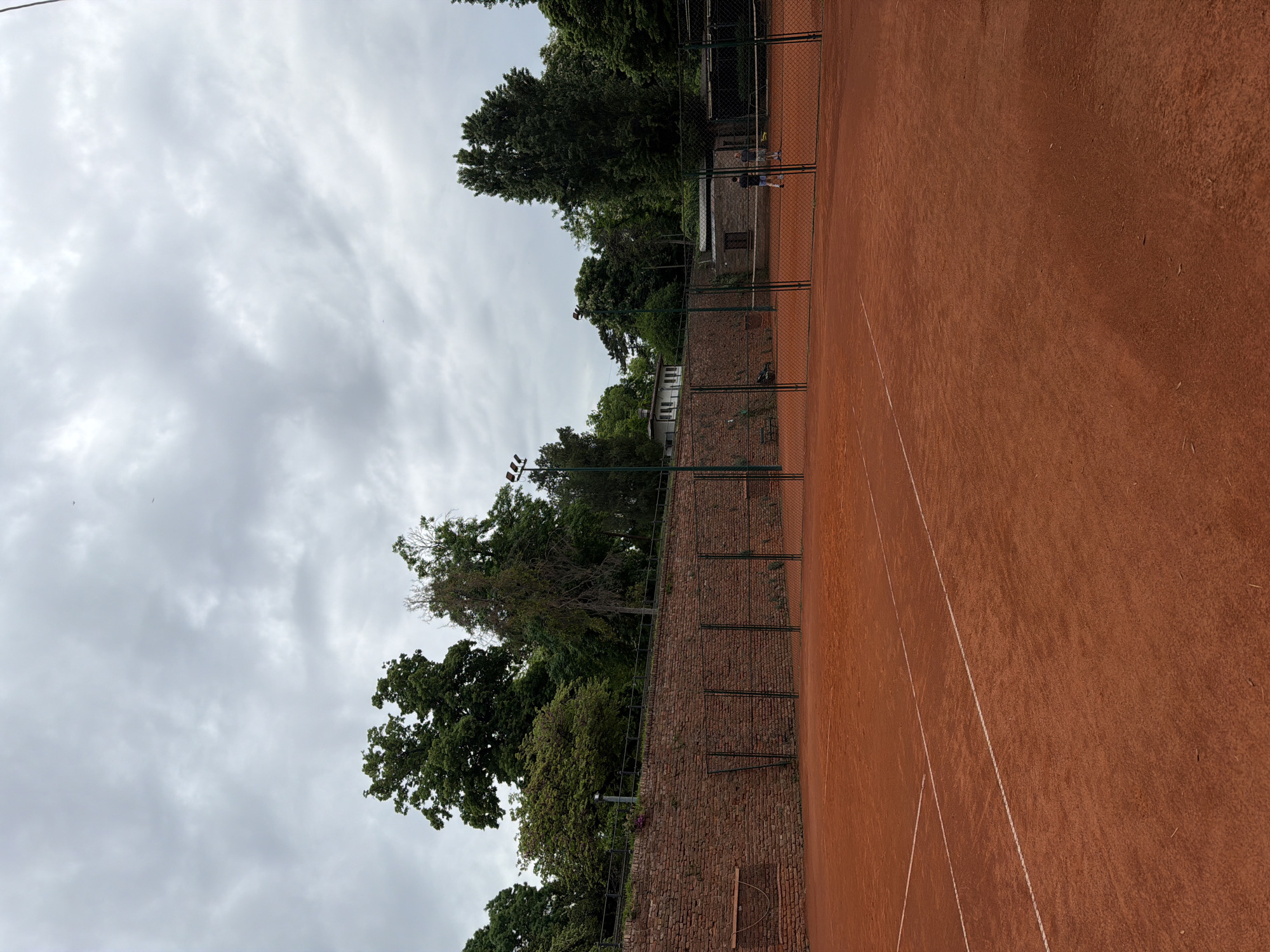 The image shows a tennis court in Belgrade, Serbia. The court is made of red clay and has white lines marking the boundaries. A brick wall with a chain-link fence runs along one side of the court, with trees visible behind the wall. There is a light post visible near the wall. In the distance, two people appear to be playing tennis. The sky is overcast with grey clouds.
