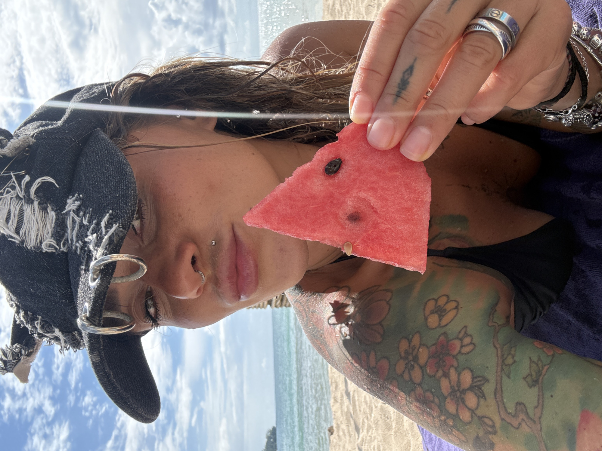 The image is a close-up, selfie-style shot of a person holding a slice of watermelon. The person is outdoors, likely on a beach, with a bright sky and a body of water visible in the background. The sun is shining, creating lens flare and highlights on the water and the person's hair.
The person is wearing a dark bandana or hat with metal grommets and distressed fabric. They have several piercings, including one on their nose and one on their cheek. They are also wearing rings on their fingers, including a band with an "X" symbol and another layered band. The person has tattoos on their arm and shoulder, featuring floral designs in various colors.
The watermelon slice is a vibrant pink, with dark seeds visible. The person's skin appears slightly sun-kissed. The overall mood of the image seems relaxed and summery, suggesting a casual outing at the beach. The time of day appears to be daytime, given the bright sunlight and clear sky, likely morning or afternoon. The location cues (beach, water, sand) combined with the provided context point to Choeng Thale, Thailand. There is no visible text in the image.