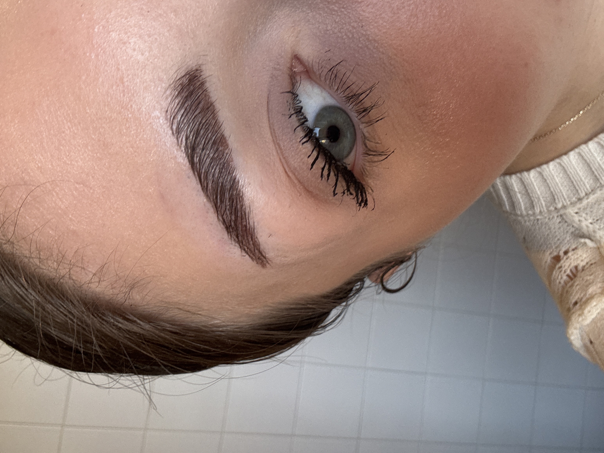 The image presents a close-up, static view of the right side of a female subject's face. The subject's right eye is open, displaying a grey-green iris and a dark pupil. Eyelashes are coated with black mascara, appearing lengthened. The eyebrow above is dark brown, well-defined, and groomed. The skin is light-toned, with visible pores and a slight redness on the cheek area. Dark brown hair is pulled back, with individual strands visible along the hairline. A section of a light-colored, ribbed knitted garment is visible on the lower right, along with a thin gold-colored chain necklace resting on the skin. The background consists of a light-colored, possibly white or pale grey, tiled wall with distinct grid lines formed by grout. The specific location in L'hospitalet De Llobregat, Spain, is not discernible from the visual information within the image.