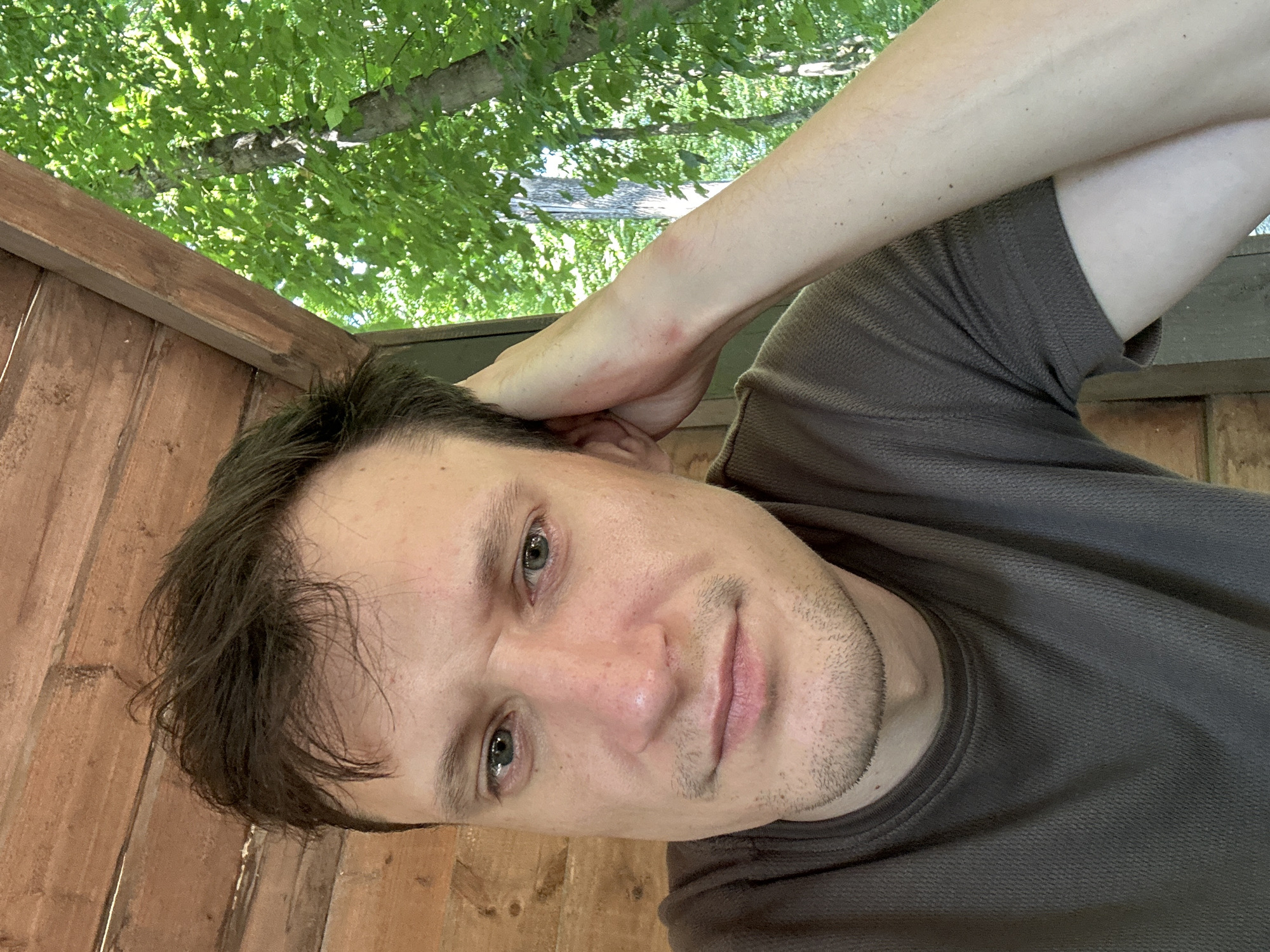 A medium-shot, low-angle selfie shows a man in his early 30s with dark hair and stubble, resting his head on his left arm. He is wearing a dark brown t-shirt. He is sitting in what appears to be a wooden structure with a wooden ceiling and wall to his left. Behind him and to the right, green foliage of trees is visible, suggesting an outdoor setting. The image seems to have been taken during the day, with natural light illuminating the scene. The man's facial expression is neutral. There is no information about the specific location in Shatava, Ukraine, provided in the image.