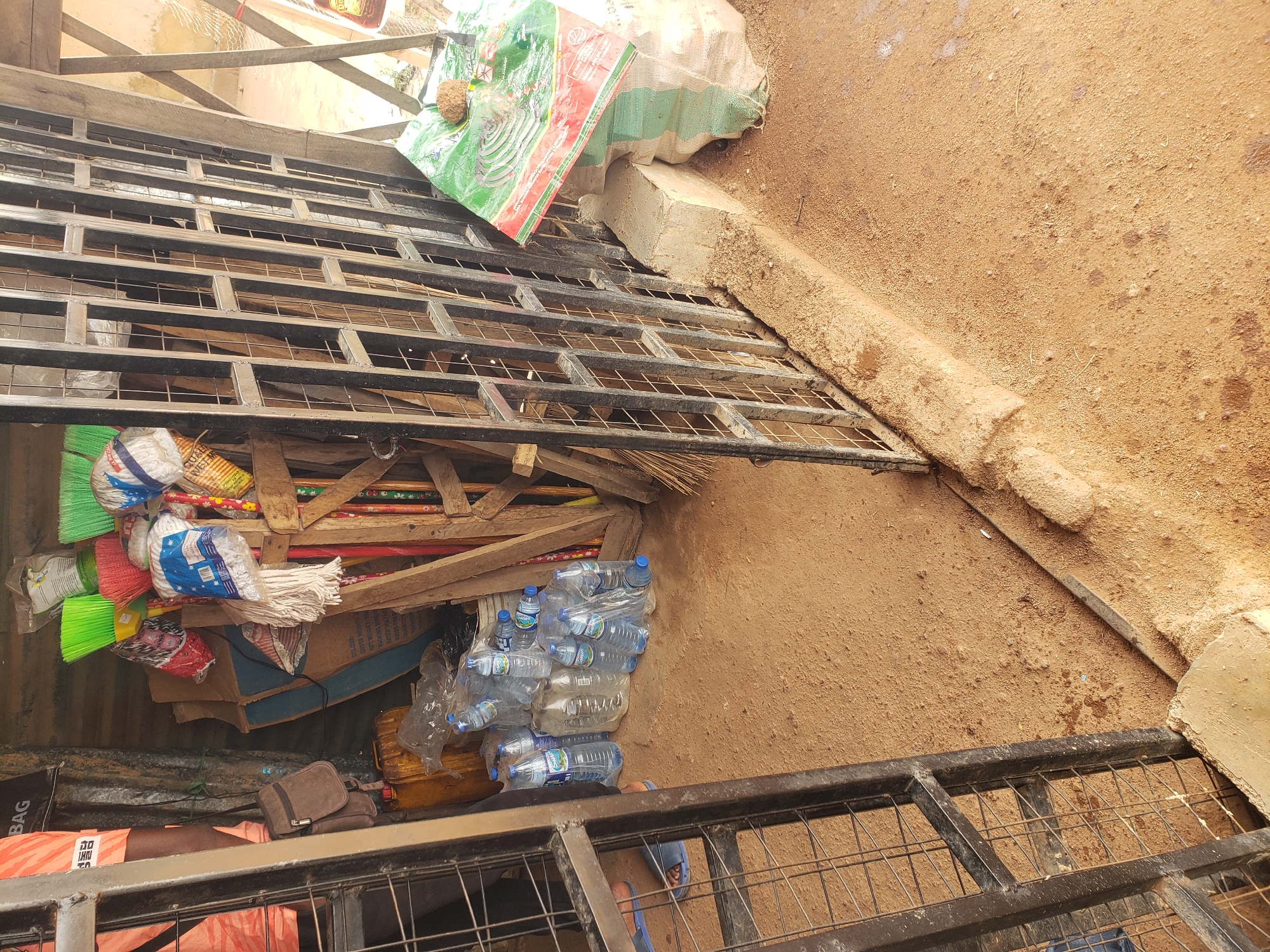 This image depicts an outdoor market stall in Bukuru, Nigeria, with a focus on household cleaning supplies. In the foreground, a black metal grate partially obscures the view into the stall. To the left, a person wearing orange shorts and a black bag slung over their shoulder is partially visible. The stall itself contains an assortment of items, including stacked plastic bottles of water, brooms with brightly colored bristles (green, red, and yellow), mops, and cardboard boxes. A large, green and red plastic bag with an image of a mosquito coil is prominently displayed on the right side of the stall, resting on a concrete block and a sack. The ground is covered in reddish-brown sand, indicative of the local terrain. The overall impression is one of a functional, somewhat cluttered, but organized retail space catering to everyday needs.