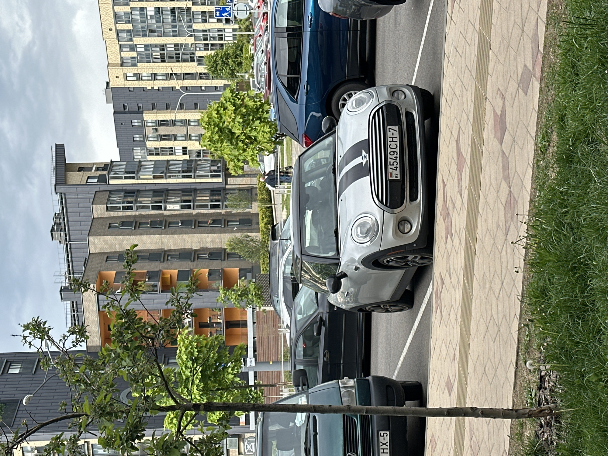 The image shows a parking area in Бараўлянскі сельскі Савет, Belarus, with several parked cars. A silver Mini Cooper with a black racing stripe and license plate "4549 CH-7" is prominently featured. A blue SUV and several other vehicles are parked nearby. Multi-story buildings with varied architectural styles line the background. A tree with green foliage is visible in the foreground, obscuring part of the view. The ground is paved with rectangular bricks and grass edges the parking area. The sky is overcast.