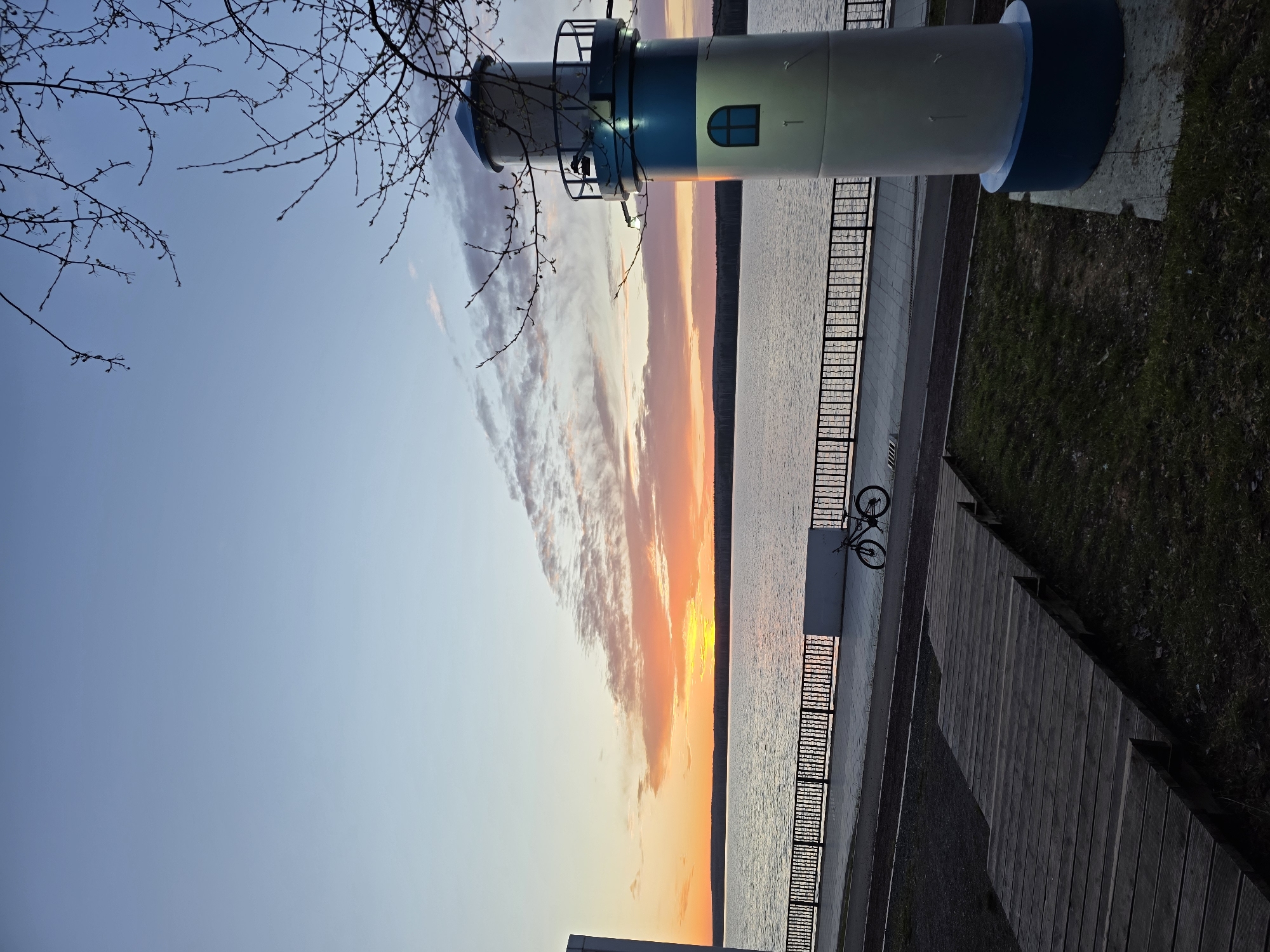 The image depicts a sunset over the Kama River in Chaikovsky, Russia, as seen from the embankment. A decorative lighthouse stands to the left of the frame, its white and blue paint illuminated by the setting sun. Bare tree branches extend into the sky above the lighthouse. The river reflects the orange and yellow hues of the sunset, while a metal fence runs along the embankment. A bicycle is leaning against a concrete structure near the fence. A wooden walkway borders the embankment on the right side, with grass visible beyond it.
