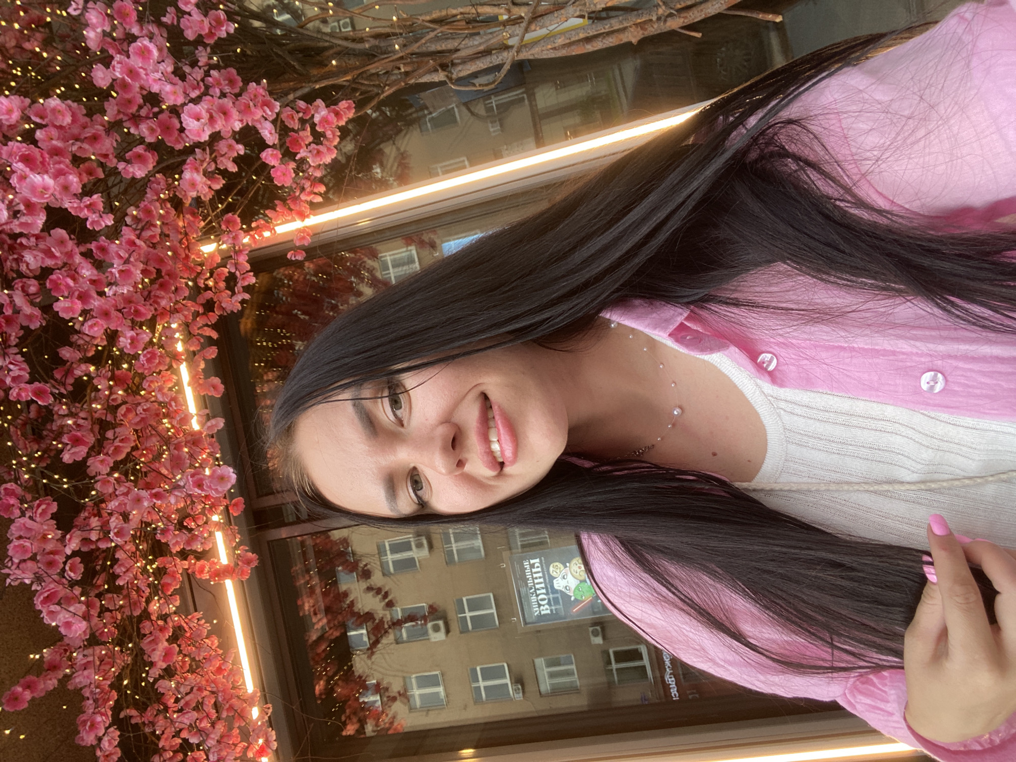 A young woman with long, dark hair is captured in a selfie, wearing a pink open shirt over a white ribbed top and a delicate pearl necklace. She has light skin, dark eyebrows, and is smiling. Her left hand, with bright pink nail polish, is holding a strand of her hair. Behind her, a building with multiple windows and air conditioning units is reflected in a large window. Above and to the left, a decorative artificial cherry blossom tree with pink flowers and small, warm-toned lights is visible, casting a gentle glow. A bright vertical light fixture is also present. The scene appears to be outdoors, possibly in front of a cafe or shop in the city of Perm, Russia, given the architectural style of the building and the text on the poster in the reflection which appears to be in Cyrillic script.