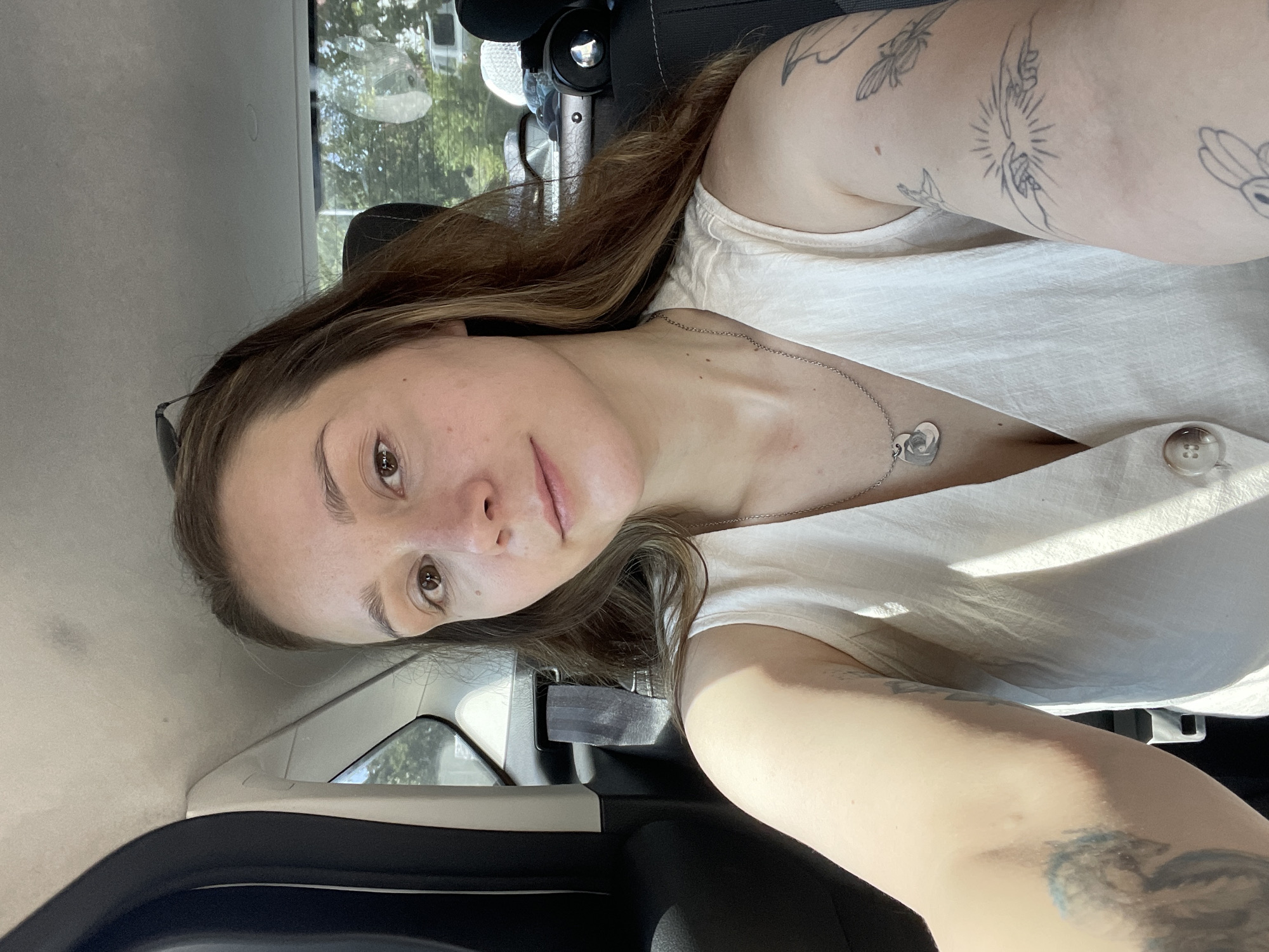 A young woman with long brown hair is taking a selfie in the back seat of a car. She is wearing a white sleeveless top and a silver necklace with a heart-shaped pendant. She has tattoos on her arms. The car appears to be moving, as evidenced by the motion blur in the background. The scene is outdoors, with trees visible through the car windows. The sun is shining, casting shadows inside the car. The location is not specified in the image, and therefore it cannot be identified as Stavropol, Russia.