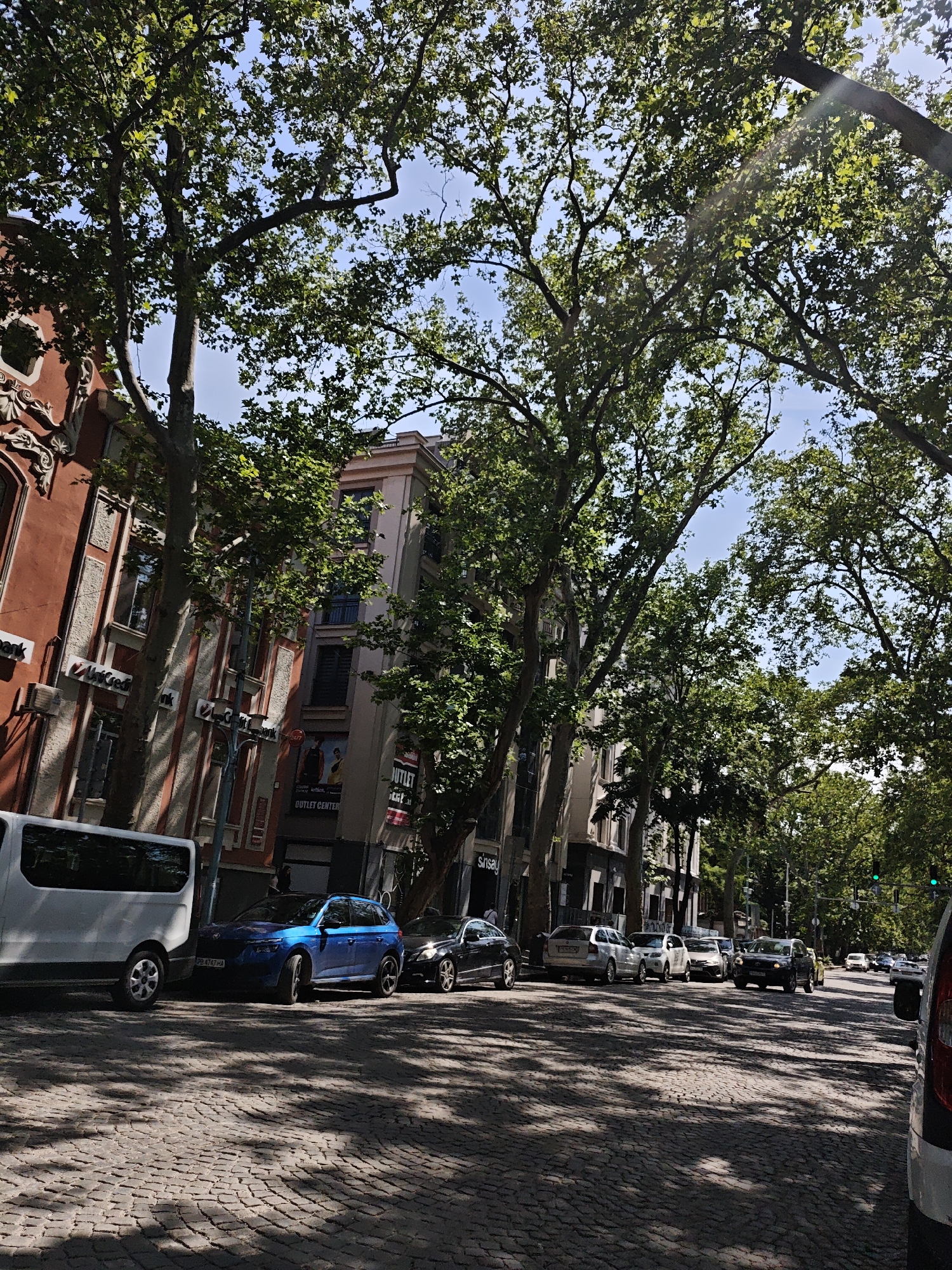 The image depicts a street scene in a city, likely in Europe due to the cobblestone road and architecture. A row of buildings lines the left side of the street, featuring red and cream colored facades with architectural details. Several trees with full green foliage are prominent, casting shadows on the street and vehicles. Parked along the curb are several cars, including a white van, a blue hatchback, and a black sedan. Further down the street, more vehicles are visible, some in motion, and traffic lights can be seen. The sky is blue with some sunlight filtering through the trees, creating a dappled light effect. The overall impression is a vibrant, sunny day in a city with mature trees lining the streets.