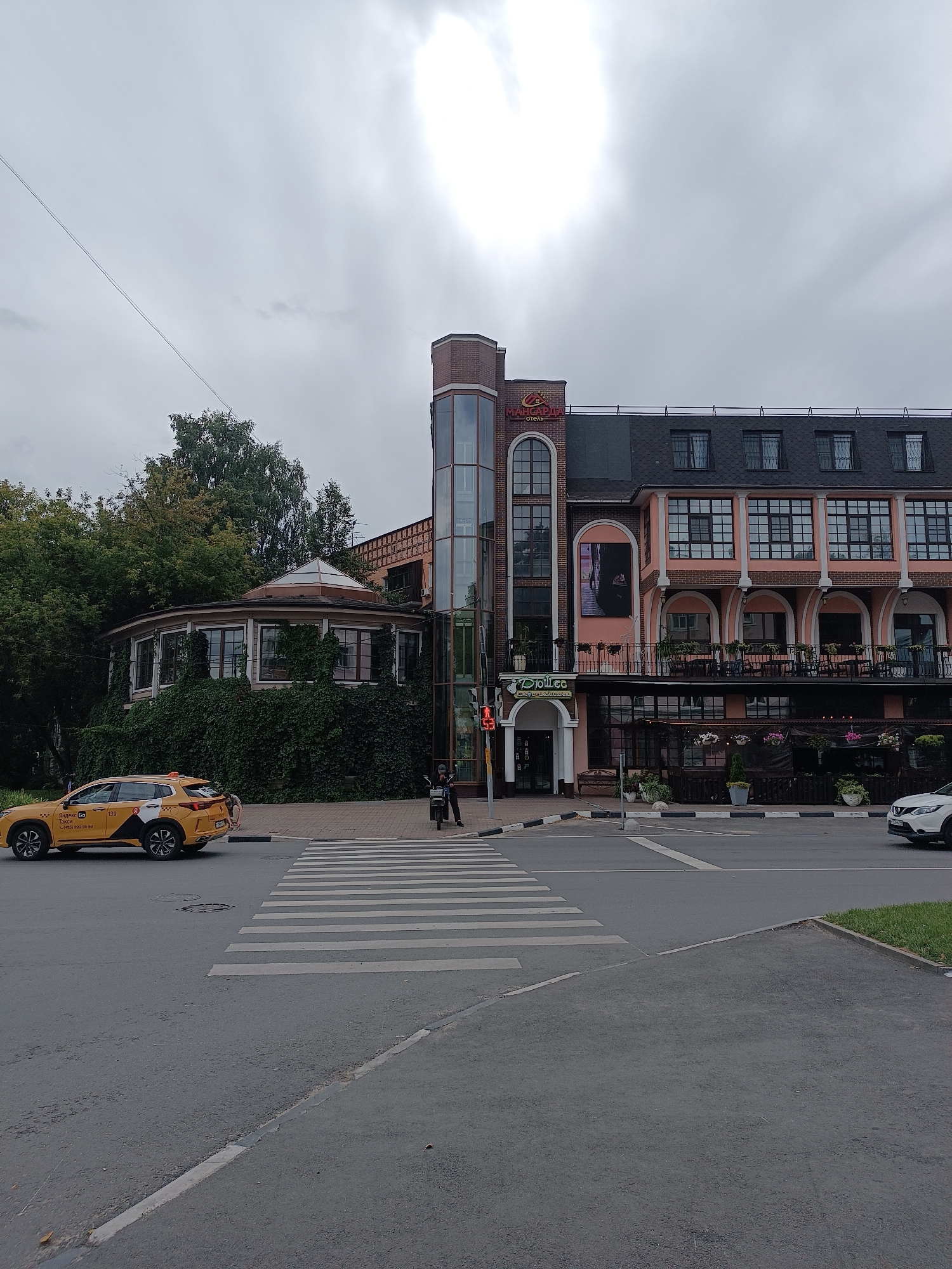 A yellow Yandex Taxi is stopped at a pedestrian crossing on a street in Lyubertsy, Russia. The taxi is a 2020 Nissan Qashqai. The building behind the taxi is the Margarita Hotel, which has a pink facade and a modern glass elevator tower. The hotel is covered in ivy on the left side. In the foreground, a white car is driving away from the pedestrian crossing. The sky is overcast.