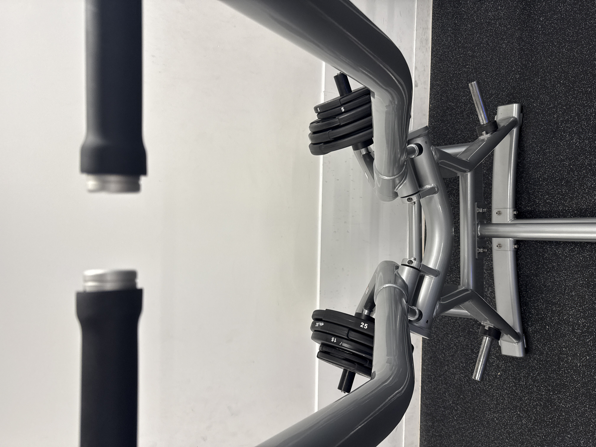 The image captures a close-up, angled view of gym equipment within an indoor fitness facility, possibly located in London, United Kingdom. No individuals are present, and no activity is occurring; the equipment appears to be either stored or set for use.
In the left portion of the frame, two black, cylindrical handles or bars are visible against a clean white wall, slightly out of focus. The right side prominently features the metallic silver and grey structure of a weight machine. Several black weight plates are stacked on a loading pin, with clearly visible white numbering indicating their denominations: "5", "10", "15", "20", and "25". Another empty loading pin is seen further to the right. The background consists of a stark white wall meeting a dark, speckled rubber gym floor, typical of a fitness environment. The composition conveys a modern and well-maintained setting.