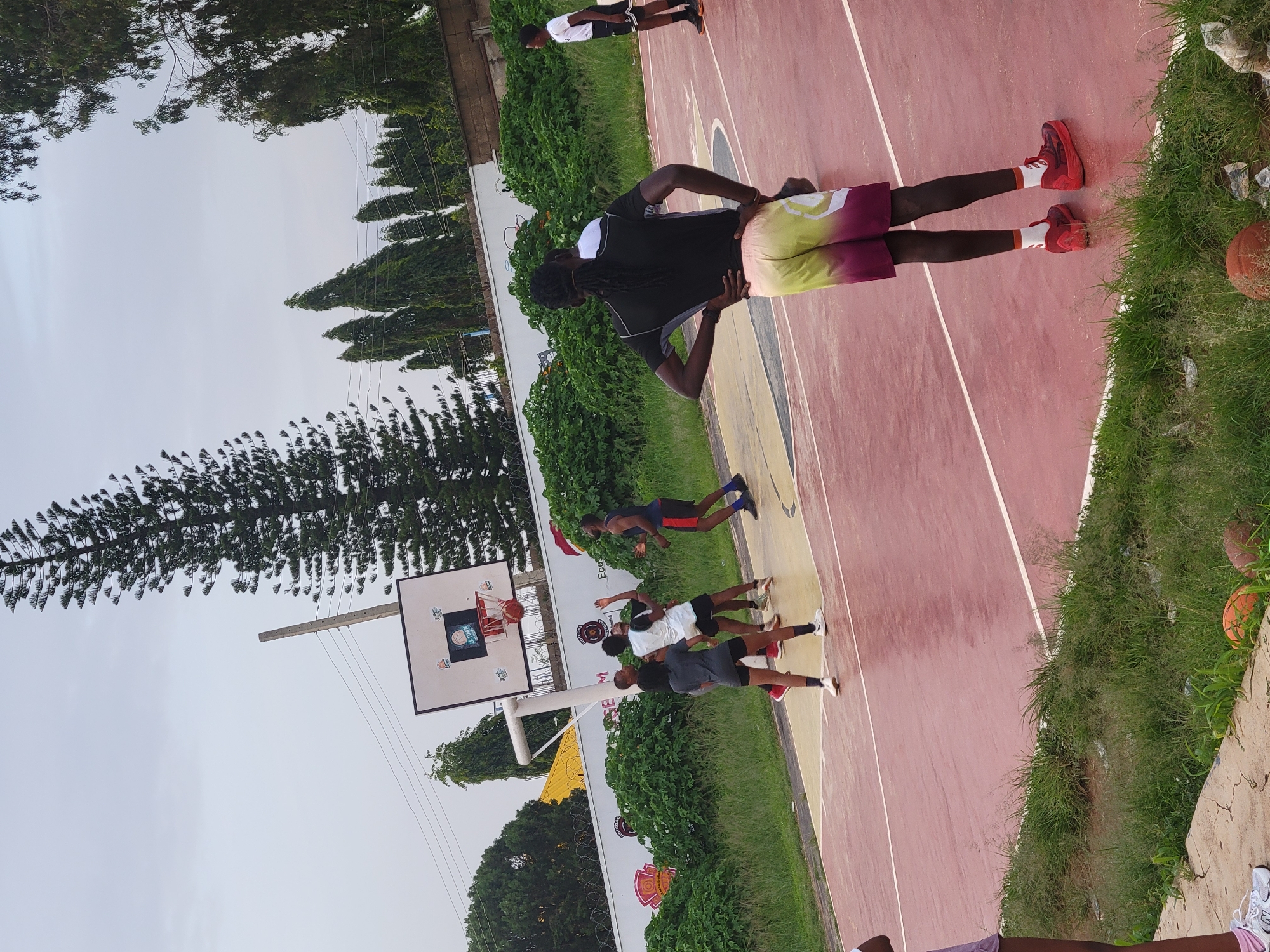 An outdoor basketball court is the setting for a game in progress. Several individuals are present on the court, engaged in play. The court surface is a muted red with yellow markings. A standard basketball hoop with a white backboard and orange rim is positioned at one end of the court. Lush green bushes and trees surround the court, and a concrete wall with graffiti can be seen in the background. The sky is overcast and a pale grey.