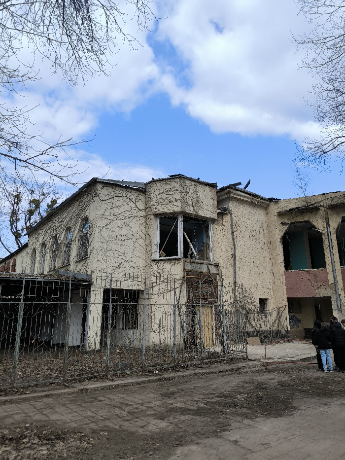 The image captures a two-story building in Vinnytsia, Ukraine, displaying severe disrepair and damage under a partly cloudy blue sky. The building's light-colored stucco exterior is extensively covered in leafless vines. The left side features arched window openings, some obscured or missing glass, above a lower section partially covered by a corrugated metal roof structure. The central portion includes a prominent bay window on the second floor with broken window frames and missing glass. Below it, a ground-level entrance with a yellow door is visible behind a metal fence. The right side of the building shows more extensive structural damage, with large, gaping openings revealing a light blue interior wall and pockmarked exterior surfaces. The roofline in this section is visibly damaged, with exposed elements. A weathered metal fence with decorative scrollwork runs across the mid-ground, separating the viewer from the building. The foreground consists of an unpaved, dirt ground mixed with dry leaves and debris, with a section of paved bricks visible to the left. Three individuals, dressed in dark attire, are positioned on the far right, near the damaged section of the building, facing towards it. Bare tree branches frame the upper perimeter of the scene.