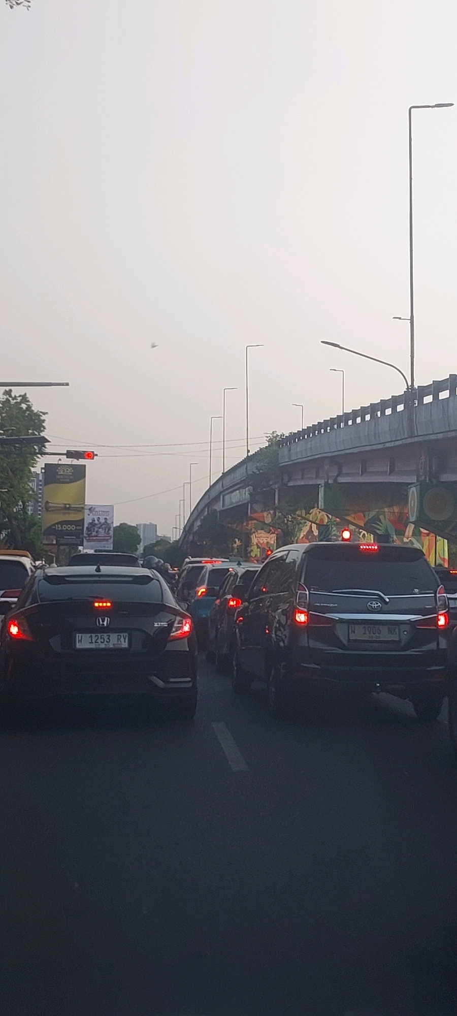 The image captures a congested traffic scene on a highway overpass in Surabaya, Indonesia. The sky is overcast with a hazy, light gray hue, suggesting a potentially polluted or foggy atmosphere. The foreground is dominated by a queue of vehicles, primarily cars, moving at a very slow pace, indicating a traffic jam.
In the center of the frame, a black Honda Civic with license plate "W 1253 RY" is prominently featured, its brake lights illuminated. To its right, a dark gray Toyota Avanza with license plate "W 1906 NX" is also visible, with its brake lights and top brake light illuminated. Other vehicles are discernible in the background, their brake lights creating a series of red glows in the dim light.
On the left side of the image, a traffic light displays a red signal, confirming the halt in traffic. Adjacent to the traffic light are billboards, one of which shows "15.000" and another a picture of what appears to be a musical performance.
The overpass structure itself, with its concrete supports and railings, occupies the upper portion of the image, curving into the distance. The underside of the overpass is adorned with vibrant graffiti, featuring colorful depictions of nature, including fruits and leaves. Streetlights with long poles are spaced along the overpass, some of them illuminated. Trees with green foliage are visible on the left, framing the scene. The overall impression is one of urban infrastructure overwhelmed by traffic congestion.