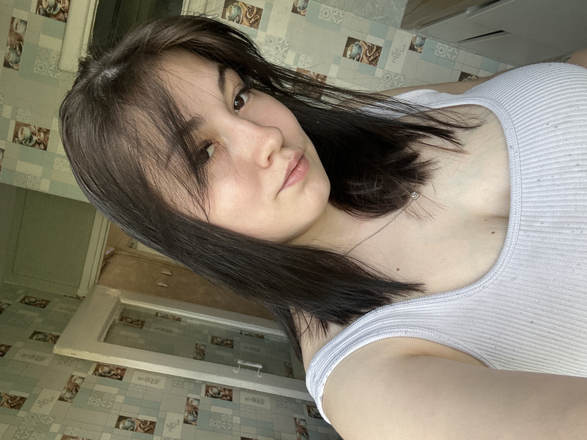 A woman with dark brown hair and fair skin is posing for a selfie. She is wearing a white ribbed tank top. A silver necklace is visible around her neck. The background consists of patterned tile walls in muted blue, beige, and brown tones. There is a window frame visible in the background with a blurry view outside. The tiles appear to be in a kitchen or bathroom. The location is Chaikovsky, Russia.