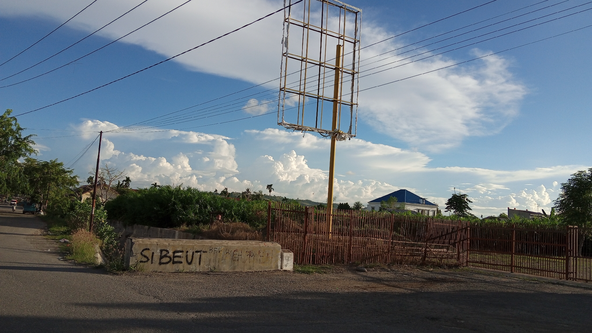 The image depicts a roadside scene in Geulangganggampong, Indonesia, under a partly cloudy sky. The foreground shows a paved road with a concrete barrier on the right side, which is graffitied with the words "SI BEUT". Behind the barrier, a rustic wooden fence encloses an area with dry vegetation. Further back, a tall, empty billboard frame stands prominently. Beyond the fence, a modern building with a blue roof is visible, surrounded by trees and other structures. The background is dominated by a vast expanse of sky filled with dramatic cumulus clouds and power lines crisscrossing the frame. A few trees line the left side of the road, and a small vehicle is seen in the distance. The overall impression is one of a developing or semi-urban area with elements of both natural landscape and man-made structures.