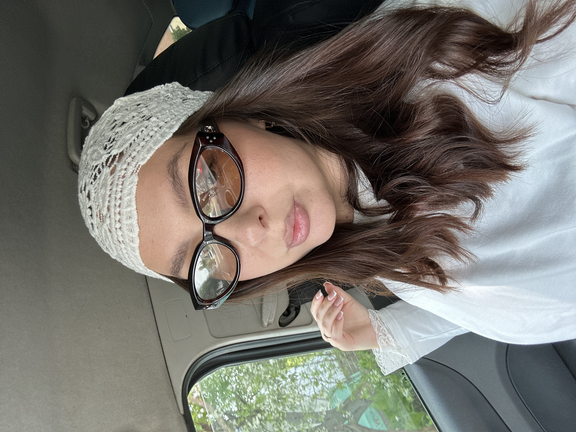 A woman with long brown hair, wearing a white lace headband and brown-rimmed glasses, sits in a vehicle in what appears to be the city of Chaikovsky, Russia. She is wearing a white shirt with lace detailing on the sleeves. The car window shows foliage and buildings outside. The interior of the car is a mix of light and dark grey.