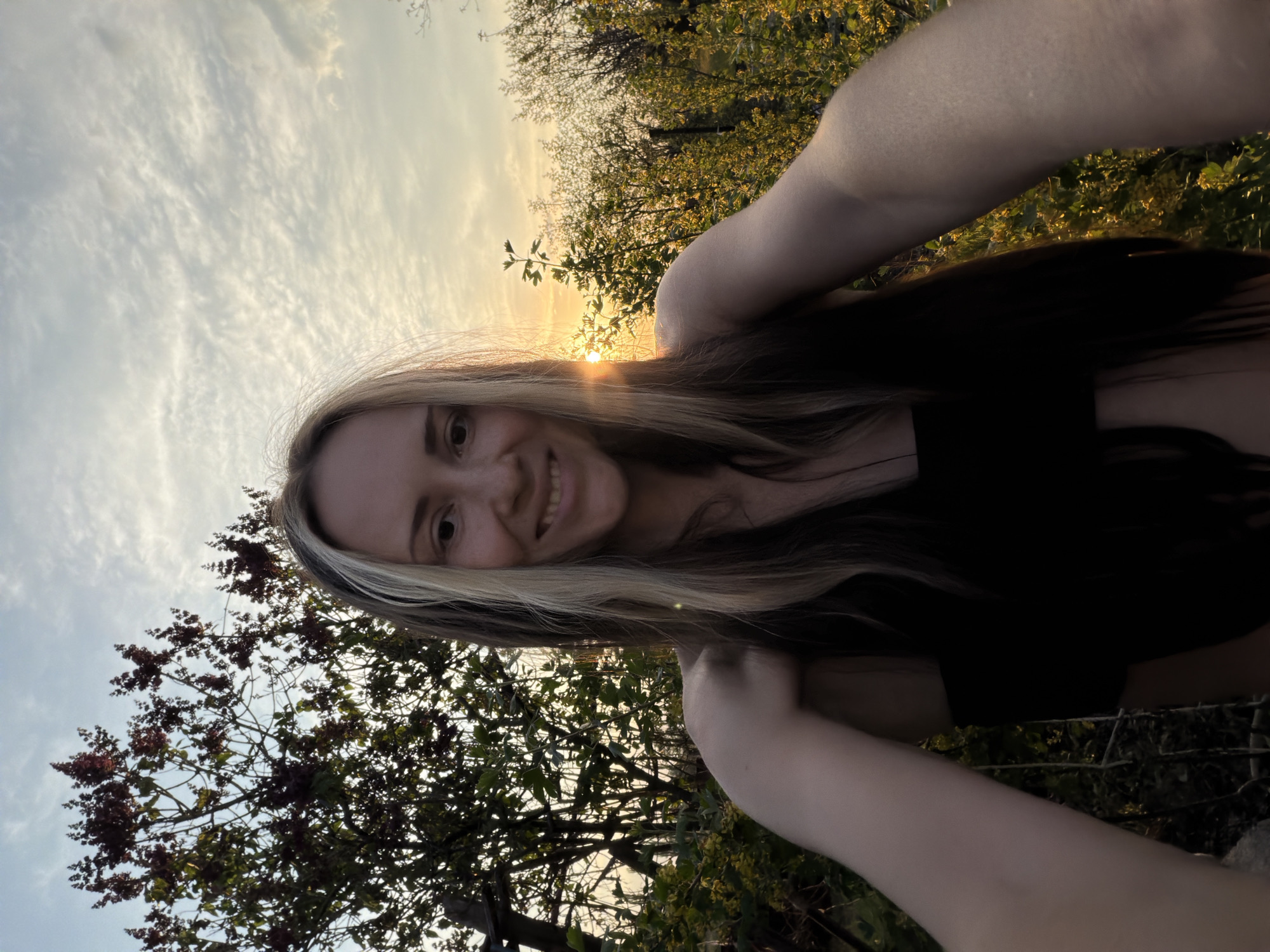 A young woman with long blonde and dark hair is taking a selfie in an outdoor setting. The background consists of trees and a sky with a sunset or sunrise. The location is suggested to be Kamyshin, Russia. The woman is wearing a black top and is smiling at the camera. Her arms are visible on either side of her face, indicating she is holding the camera herself. The lighting suggests the photo was taken during golden hour.
