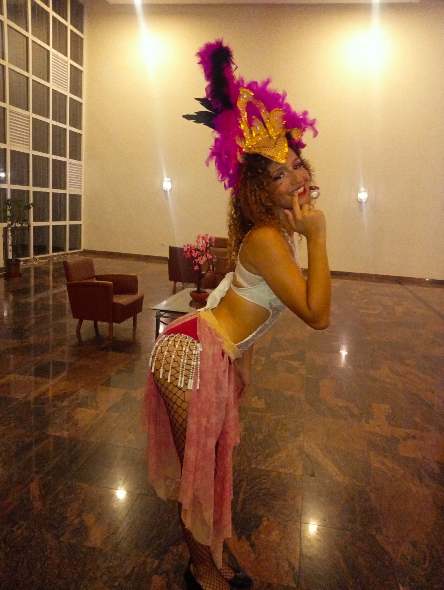 The image shows a woman in a festive costume posing in what appears to be a hotel lobby. The woman is wearing a white top, a red and pink skirt with beaded fringe, and fishnet stockings with black shoes. On her head, she has a large headdress decorated with purple feathers and a gold crown. She is slightly bent over, with her left hand raised to her chin, smiling at the camera.
The lobby has brown marbled flooring, with seating arrangements including brown armchairs and a glass-topped table with a potted plant in the background. The walls are beige with sconce lighting, and a window with a grid pattern is visible on the left. The lighting is generally warm, with reflections visible on the floor.
The location is Pedro González, Venezuela.
