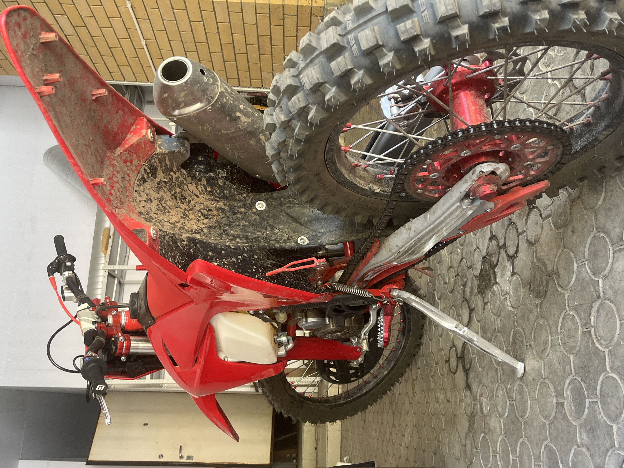 The image shows a red dirt bike, viewed from a low angle, angled slightly towards the rear. The bike is standing on a silver kickstand on a patterned concrete floor. The rear tire is a knobby off-road type. The exhaust pipe is silver and positioned close to the rear tire. The bike is splattered with mud. The background consists of beige wall tiles and a metal storage rack. There is no evidence to determine the location of this image is in the city of Ореховка, Russia.