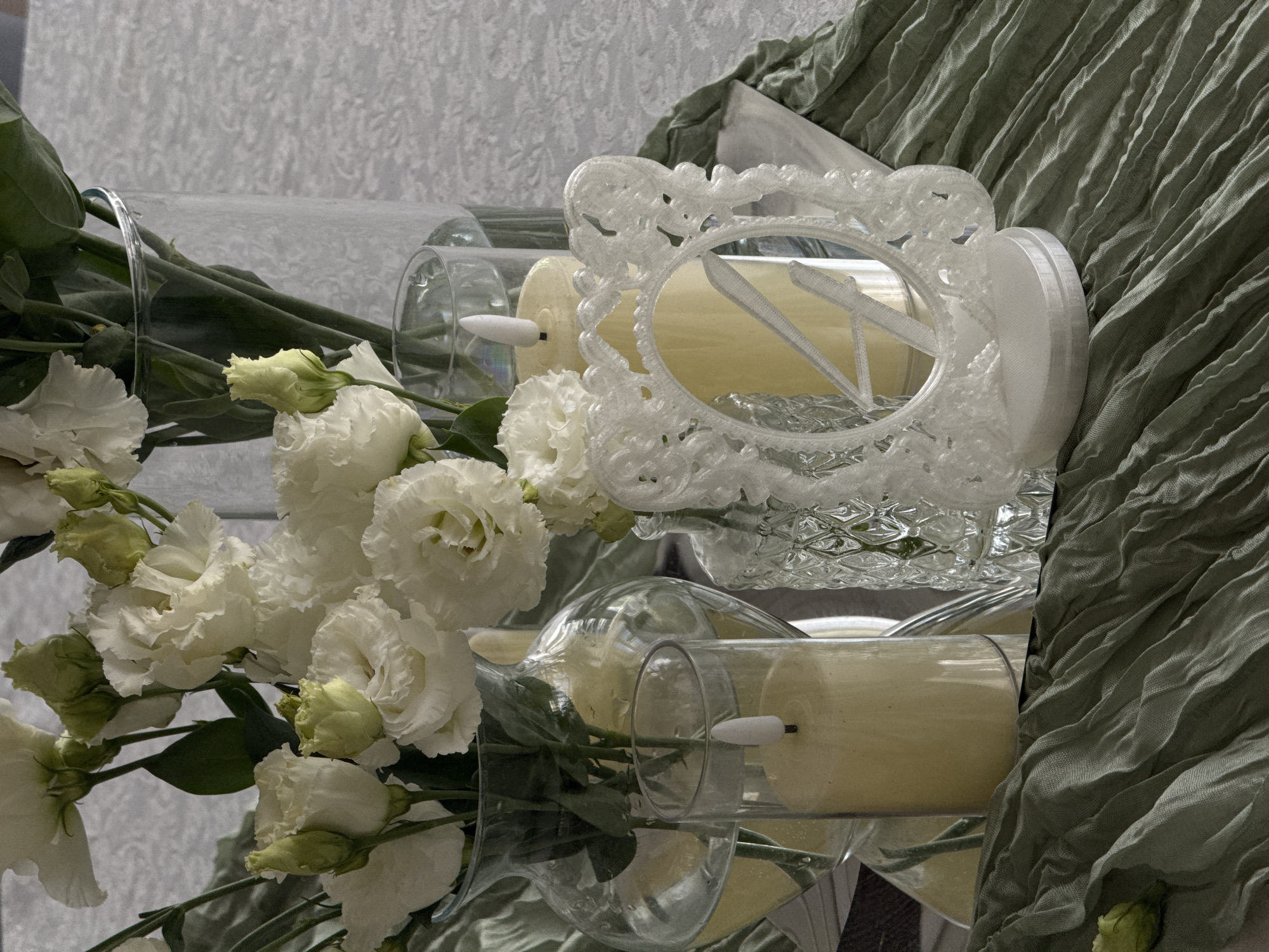 The image depicts a close-up, still-life arrangement on a table, likely for an event or as a decorative centerpiece. The scene is dominated by white flowers, possibly Eustoma (Lisianthus), arranged in various glass vases of different shapes and sizes. Some of the flowers are fully bloomed, while others are still in bud.
In the foreground and mid-ground, there are several candles. Two are visible in clear glass cylinders. One appears to be a real candle, partially lit, emitting a soft glow. The other, larger candle, is a pale yellow and has an artificial flame.
A distinctive element is a white, ornate, oval-shaped mirror with a decorative frame, positioned in front of the candles and flowers. The mirror reflects a large, light-colored letter "N" or "K". The mirror is supported by a white base.
The arrangement is set against a light-colored tablecloth with a subtle textured pattern, possibly damask or brocade. A wrinkled, sage-green fabric runner is draped across the table, adding a contrasting color and texture. A single, closed green bud or flower is visible at the bottom left of the frame, resting on the green runner.
The lighting is soft and diffused, creating a serene and elegant atmosphere. There are no people visible in the image. The scene is carefully composed and suggests a focus on aesthetic appeal, possibly for a wedding, formal dinner, or other celebratory occasion. The location in Votkinsk, Russia is not discernible from the visual elements of the image itself.