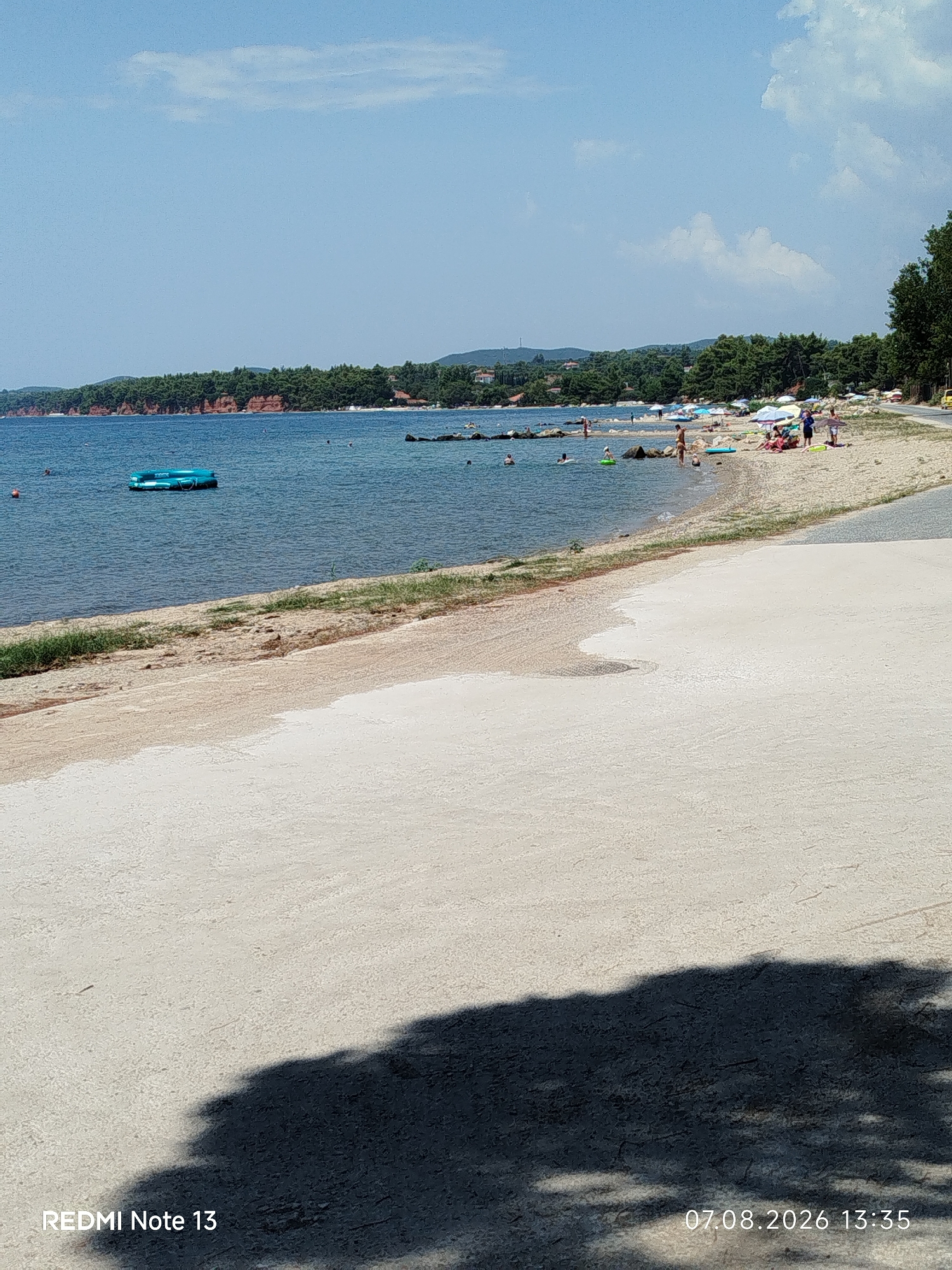 The image depicts a sunny day at a beach in the Sithonia Municipal Unit, Greece. The foreground is a paved area with a shadow of foliage cast upon it. Beyond this, a sandy beach slopes down to the sea. The sea is calm and blue, with several people swimming and relaxing in the water. A blue inflatable raft is visible floating in the sea. Along the shoreline, beach umbrellas and sunbathers are present. The background features a dense green forest line with some houses interspersed. Further in the distance, rolling hills are visible under a bright blue sky with scattered white clouds. The bottom right corner of the image contains a timestamp: "07.08.2026 13:35" and the bottom left corner displays "REDMI Note 13".