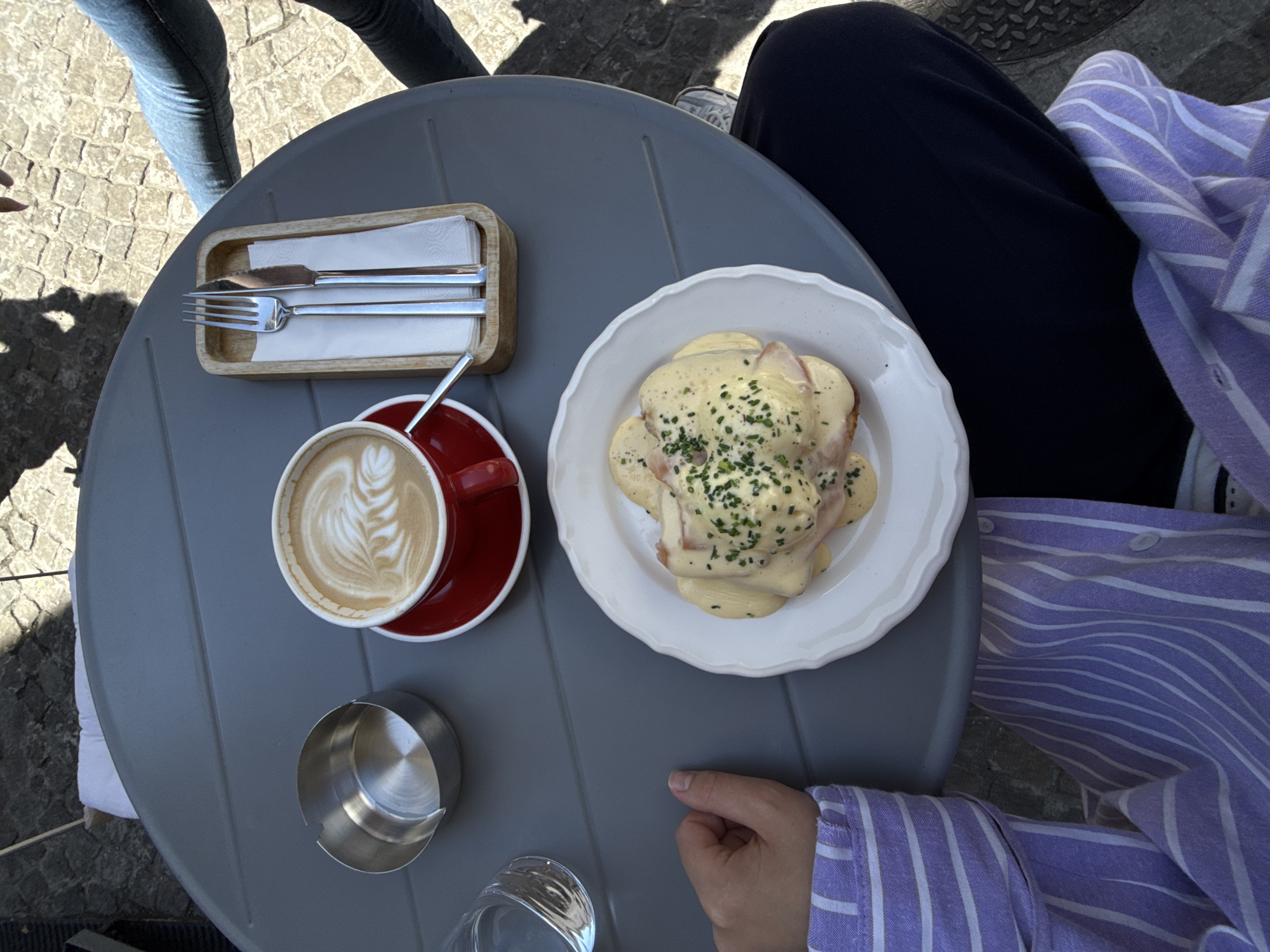A person wearing a purple and white striped shirt is sitting at a gray outdoor cafe table. On the table are a cup of coffee with latte art, a white plate with eggs benedict, a metal ashtray, a glass of water, and a wooden holder with a fork and knife. The background shows cobblestone pavement and the legs of another person wearing jeans and sneakers.