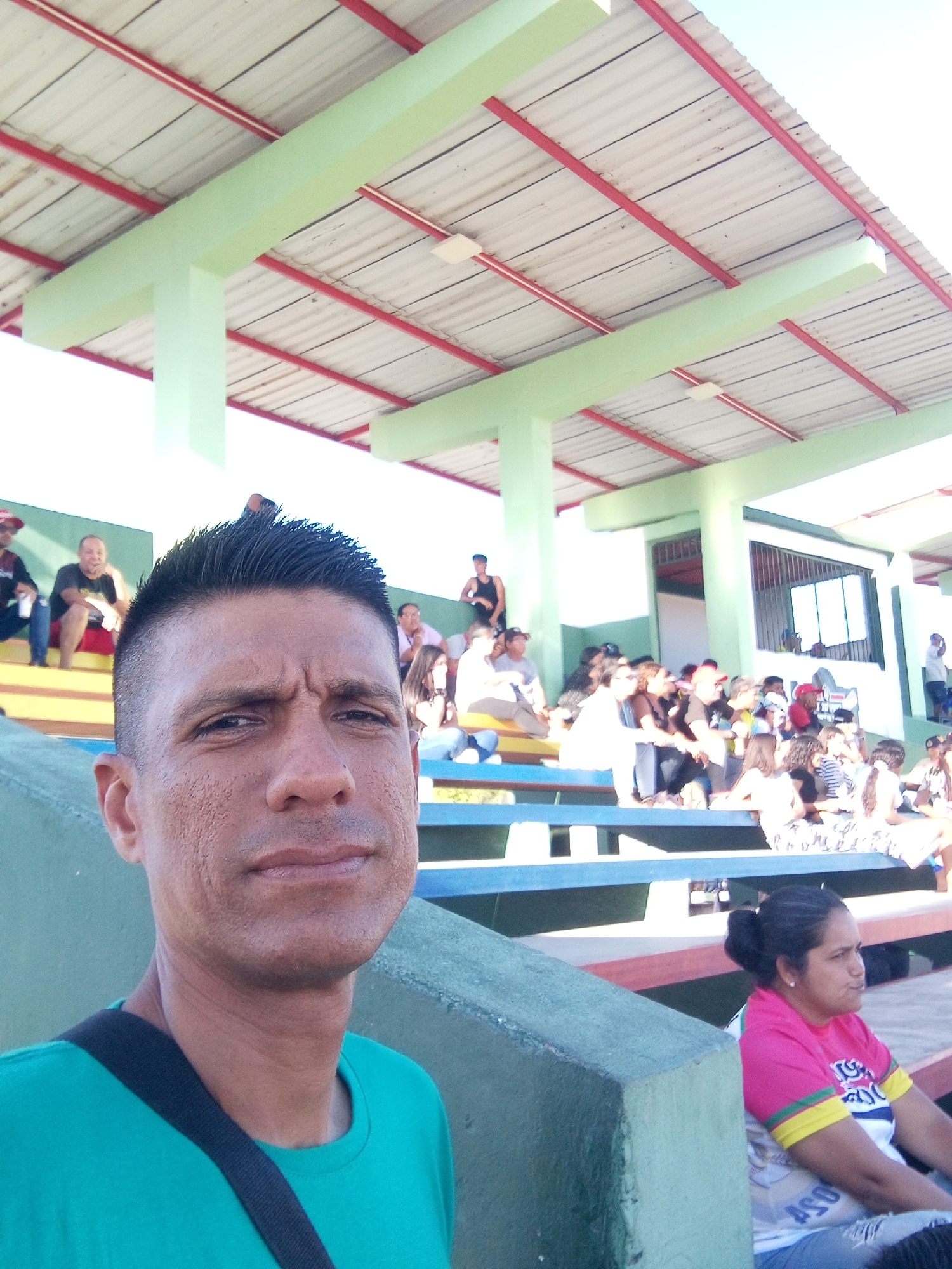 The image shows a man taking a selfie in front of a crowd of people seated in stadium bleachers. The location is likely a sports stadium in Juan Griego, Venezuela. The man is wearing a green shirt with a black strap across his chest. He has short, dark hair. The bleachers are blue, red, and green. A woman with dark hair pulled back is sitting in the front row to the right. The stadium has a light green and red roof with white corrugated metal. There are other people on the bleachers.