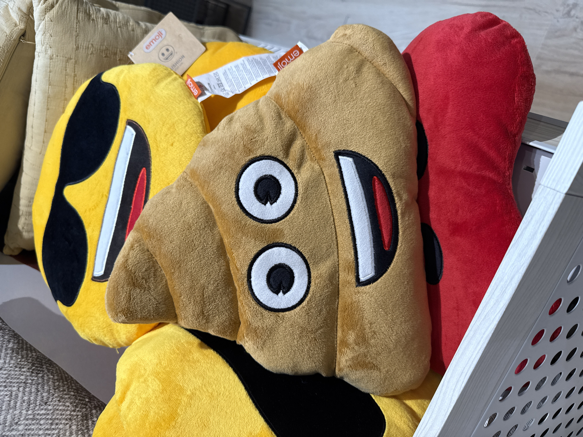 The image shows a close-up of three emoji plush cushions in a display, likely for sale. The foremost cushion is a brown "pile of poo" emoji. Behind it to the left is a yellow "sunglasses" emoji cushion. To the right of the brown cushion is a red cushion shaped like a long vertical pill. Tags are visible on some of the cushions indicating they are official "emoji" merchandise. The cushions are likely located in a retail store in Stara Zagora, Bulgaria.