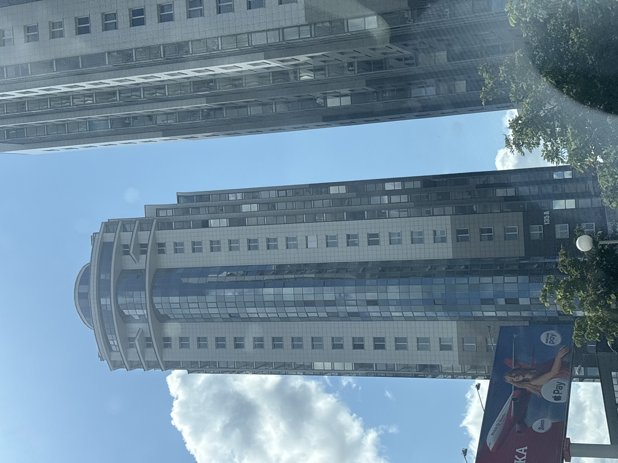 In the city of Minsk, Belarus, this photograph captures two tall, modern apartment buildings against a bright blue sky with scattered white clouds. The building on the left is a curved, glass-fronted skyscraper with a lighter colored facade. It features numerous windows and several protruding balconies. The building on the right is a more rectangular, grey structure also with many windows. In the foreground, partially visible, is a large billboard with an advertisement for "n bank" featuring an airplane and a smiling woman. The billboard also displays logos for "Swoop," "Pay," and "Samsung Pay," as well as an Apple Pay logo. Lush green trees are visible at the bottom of the image, partially obscuring the base of the buildings and the billboard.
