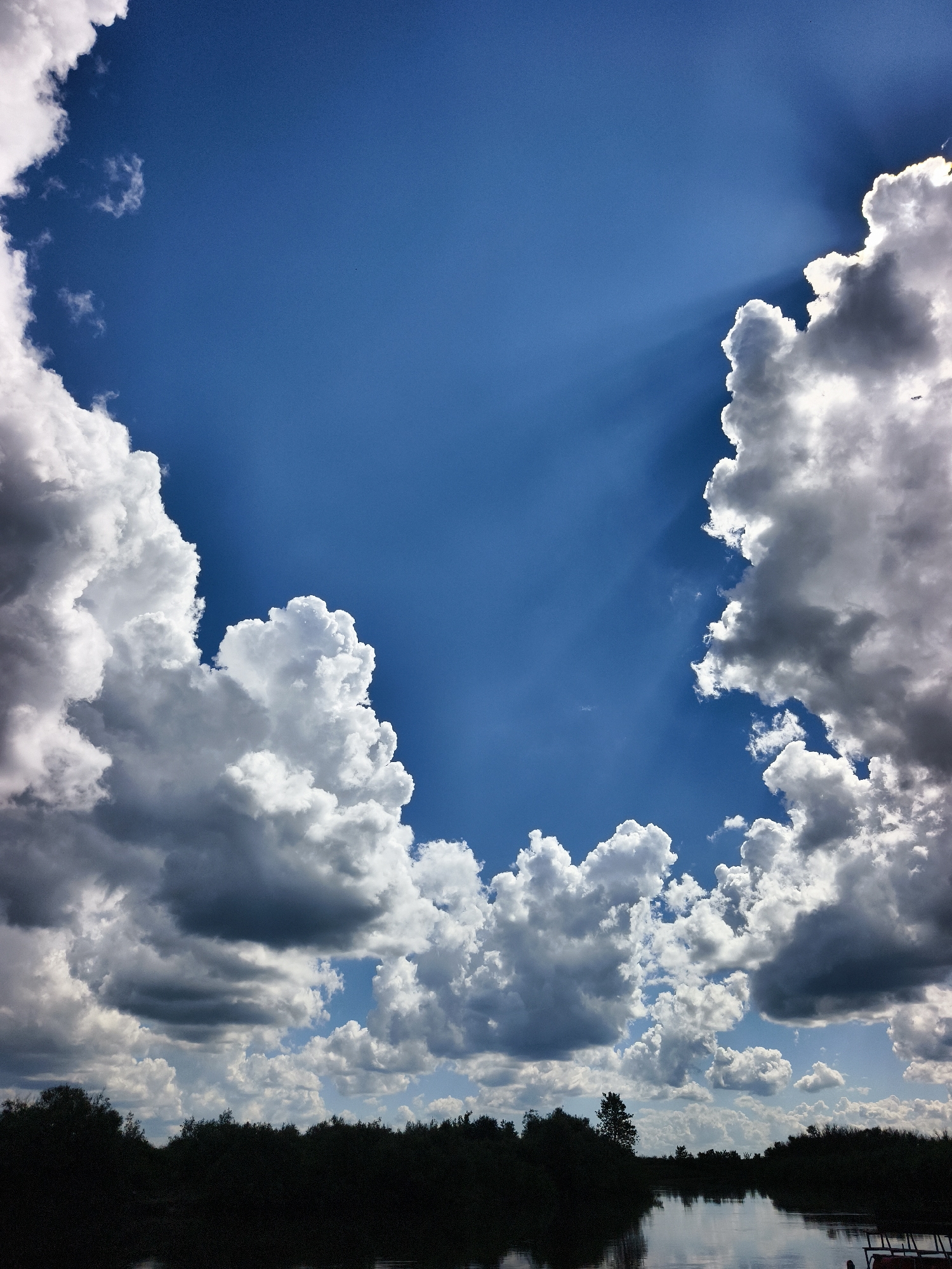 The image depicts a wide shot of a body of water with a dark, silhouetted treeline along its banks. Above the treeline, a dramatic sky is filled with large, cumulus clouds that range from bright white where the sun hits them to dark gray in their shadowed undersides. Sunbeams pierce through gaps in the clouds, creating a dramatic lighting effect. The water in the foreground reflects the sky and the clouds. The scene is likely a natural landscape, possibly a river or lake, with dense vegetation forming the horizon. The location in the city of Онохино, Russia, suggests a potentially rural or semi-rural setting within that area, characterized by natural waterways and foliage. There are no people or discernible actions captured in this still image.