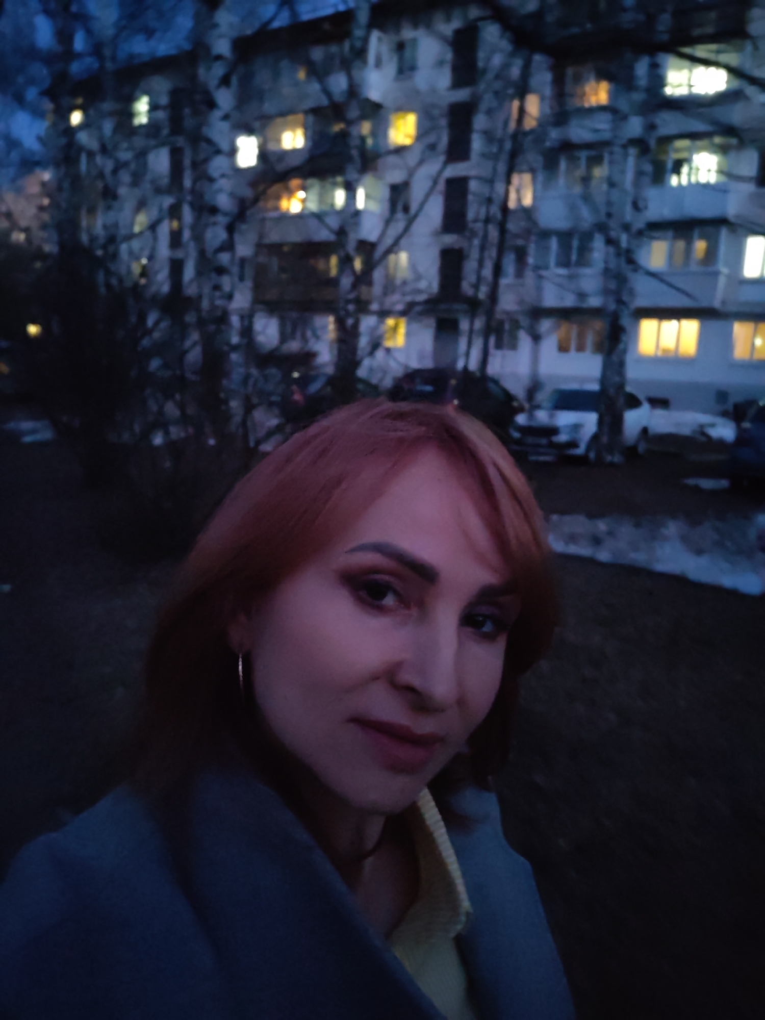 This evening selfie captures a woman in the foreground against an urban residential backdrop in Chaikovsky, Russia. The woman, with reddish-brown hair styled with bangs and wearing a light blue or grey coat over a yellow top, looks directly at the camera with a neutral to slightly pleasant expression. A silver hoop earring is visible on her right ear.
The background is dominated by a multi-story apartment building with numerous windows, many of which are illuminated with warm yellow and bright white lights, indicating evening or nighttime. Bare-branched trees, appearing to be birches, stand between the camera and the building, partially obscuring its facade. Several parked cars are visible in the mid-ground, and the dark ground shows patches that could be remnants of snow or ice, suggesting cool weather. The sky is a deep twilight blue. No visible text is present in the image.