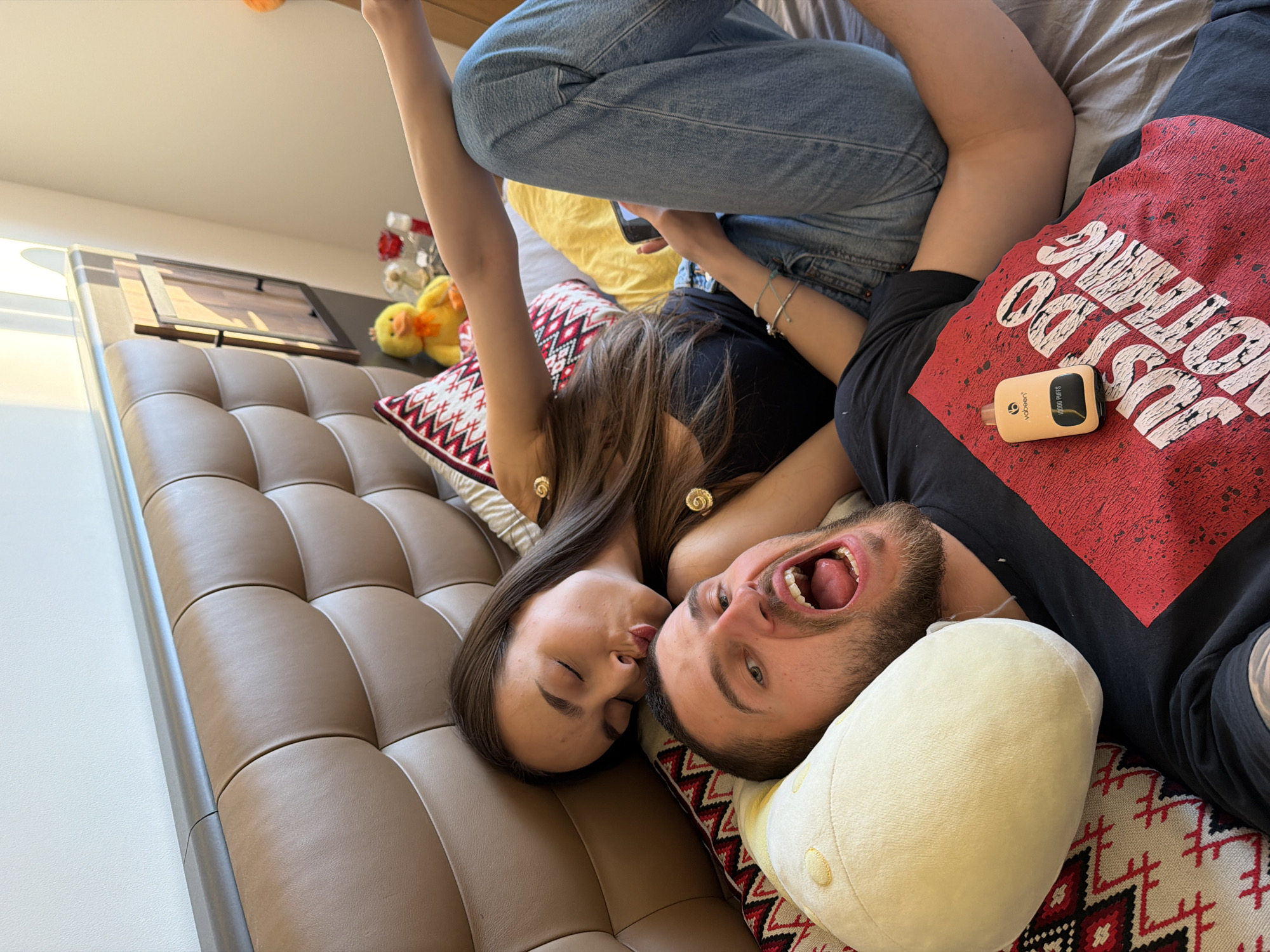 A horizontally-oriented medium shot depicts a man and a woman lying on a bed or couch. The woman, with long brown hair and wearing a black top, has her eyes closed and her lips pursed as if to kiss the man. The man, with short brown hair and a beard, has his mouth open and his tongue protruding. He is wearing a black t-shirt with a red design. A yellow vape device sits on the man's chest. Both are lying on decorative pillows. The woman's legs, clad in blue jeans, are raised in the air. The background includes a gray bed or couch, a yellow pillow, a wall, a picture frame, and yellow stuffed ducklings. The location is unspecified, but the city is Sofia, Bulgaria.